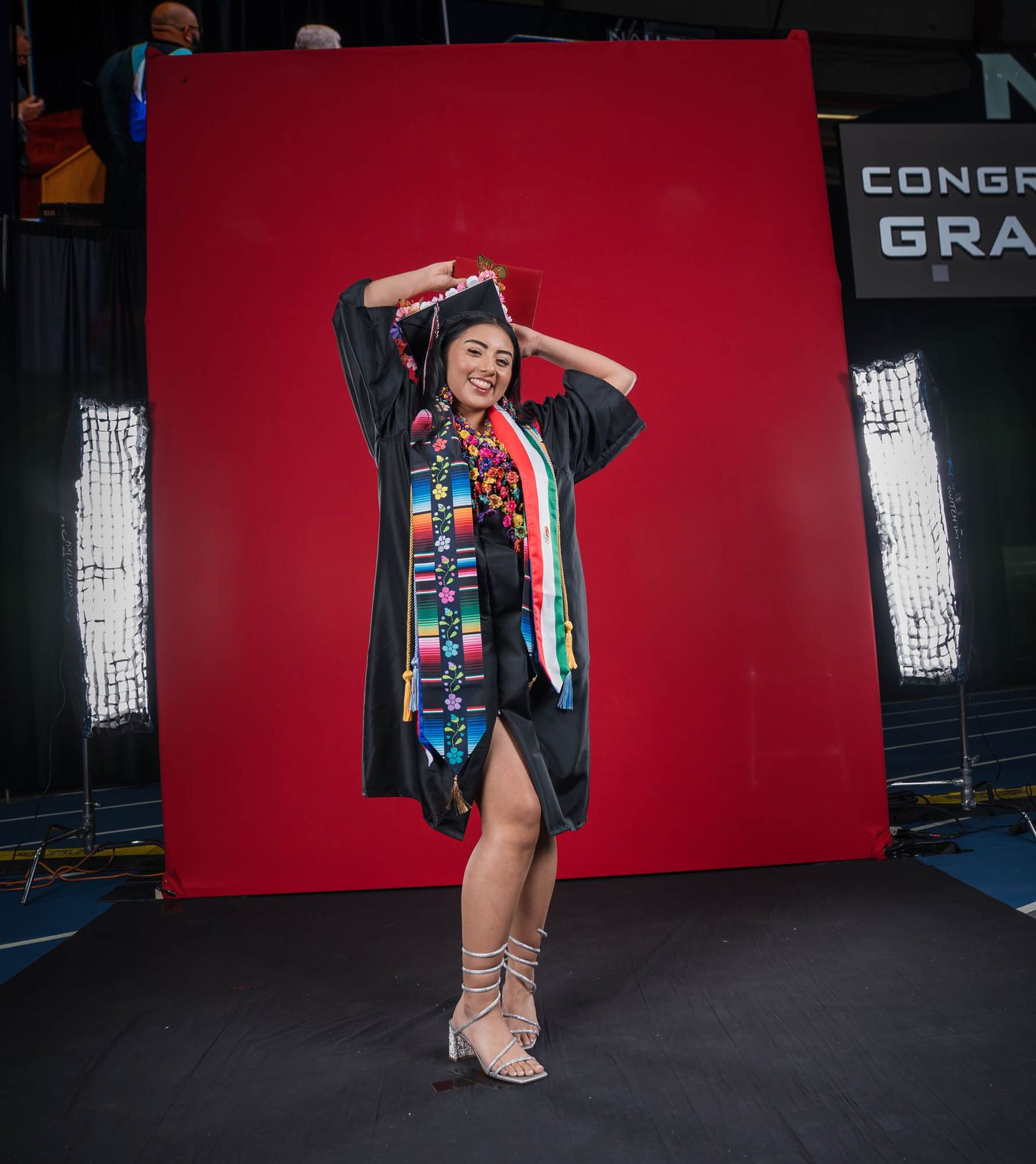 2025 CHS graduate in black cap and gown poses energetically against a bright red backdrop during commencement photo session.