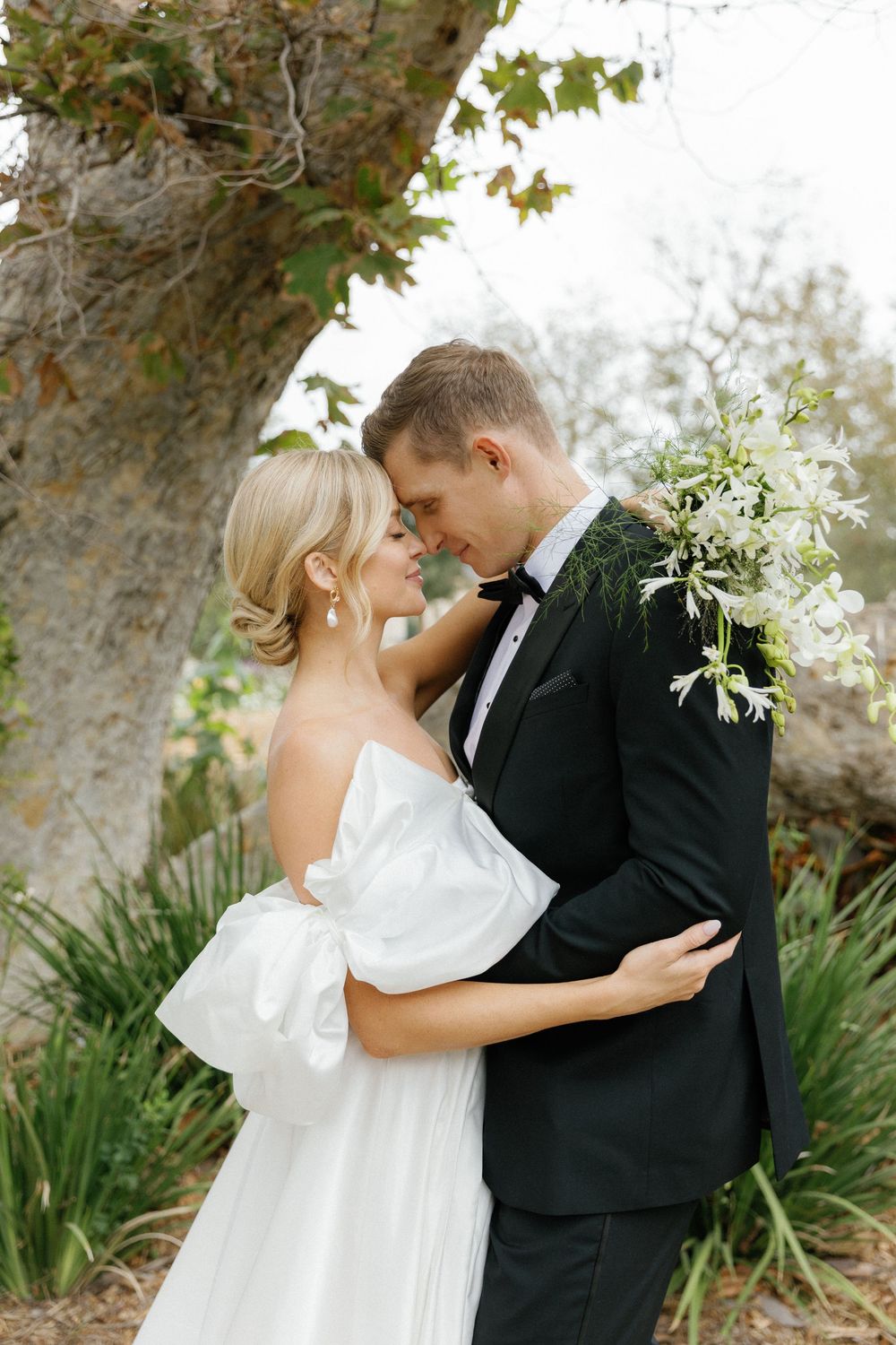 A romantic moment between newlyweds embracing outdoors with a large tree and foliage in the background.