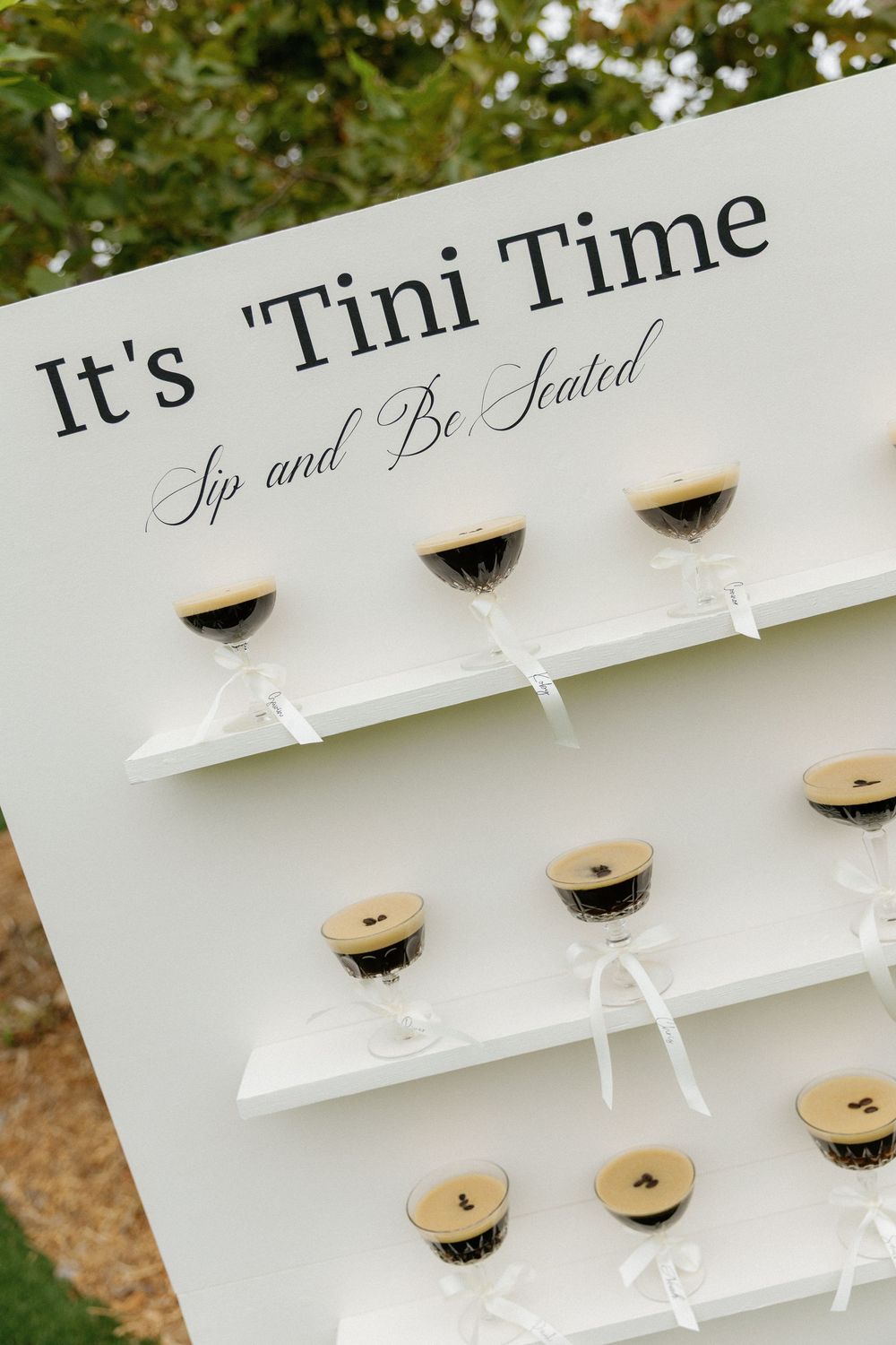 A white display wall with 'It's Tini Time' text and shelves holding mini martini glass party favors.