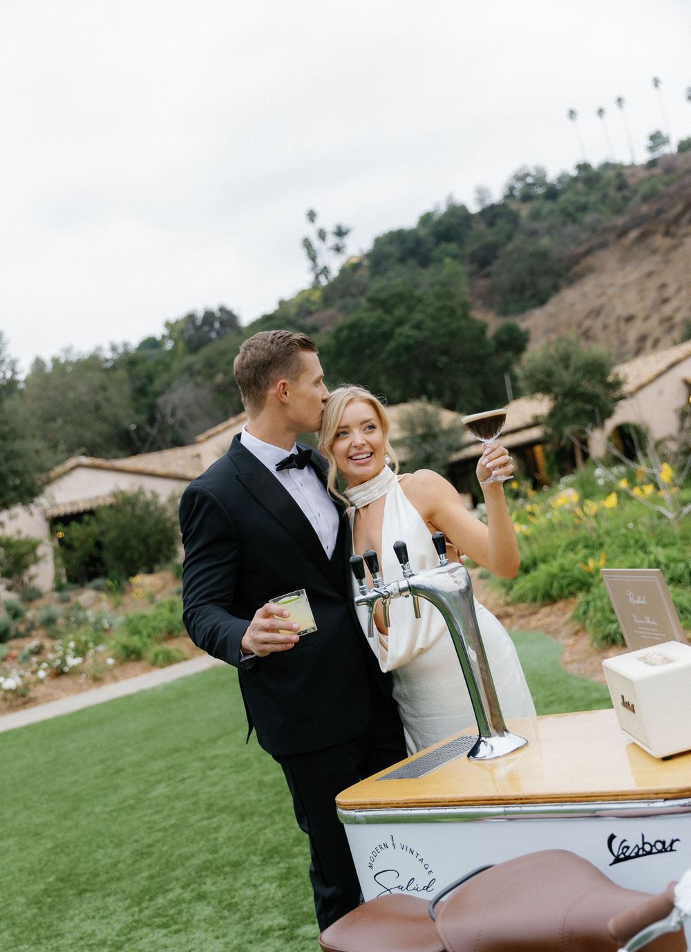 A couple enjoying wine at an outdoor party bar cart with a Vespa scooter design in a scenic garden setting.