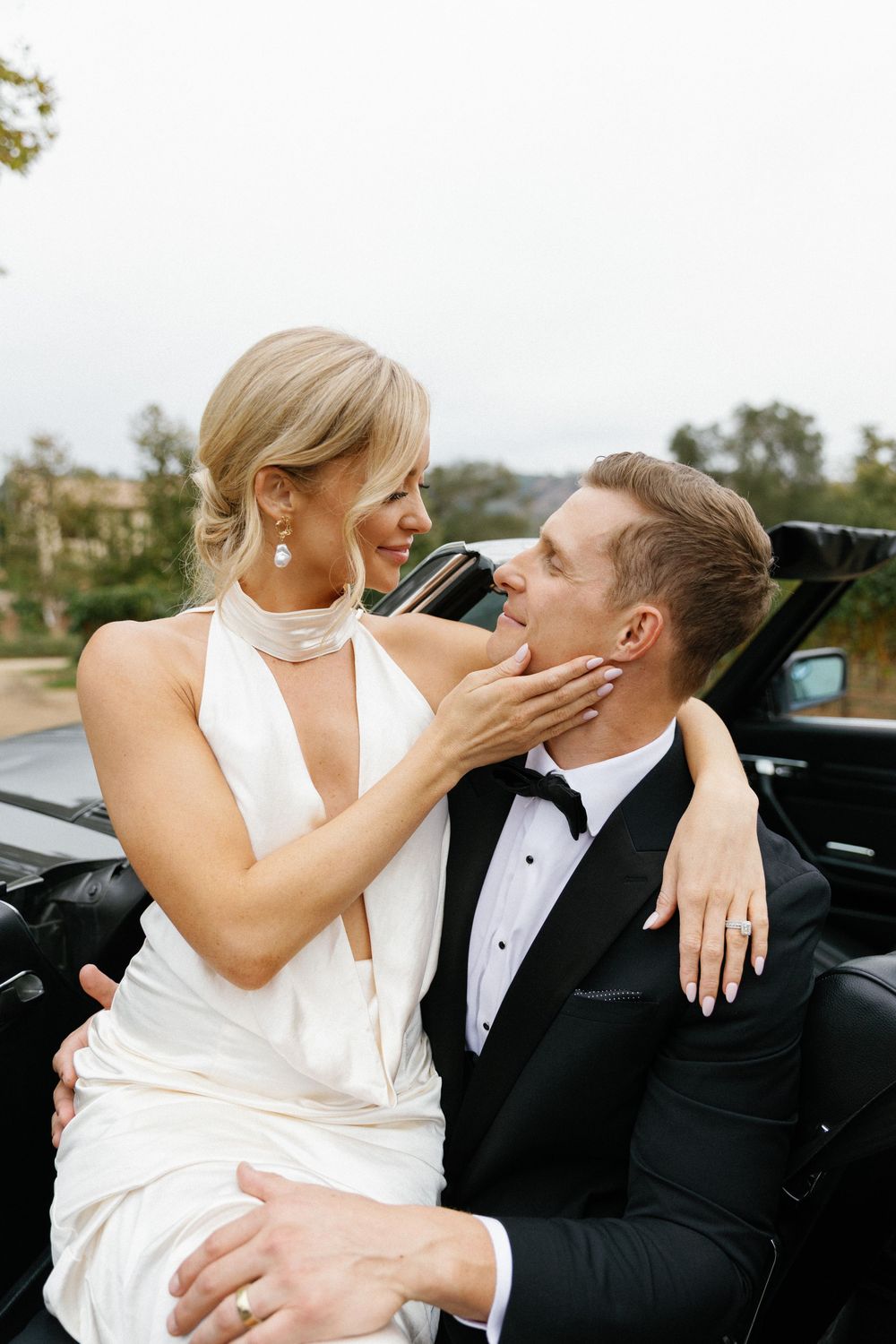 A newlywed couple share an intimate moment beside their car, with the bride in white and groom in a black tuxedo.