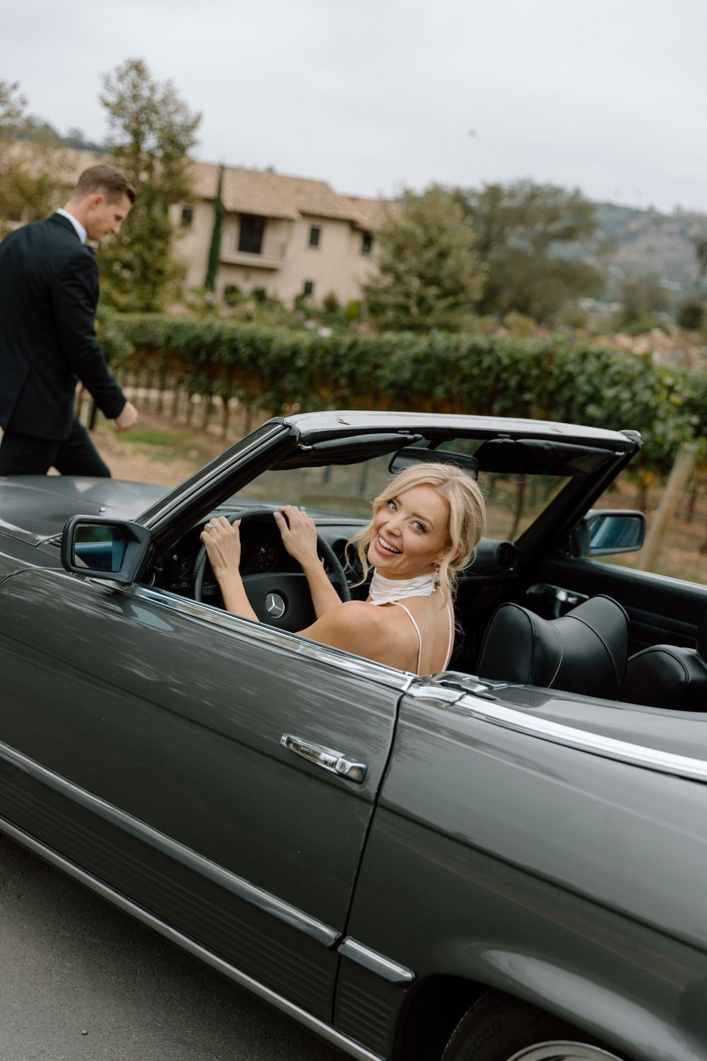 Smiling blonde bride in a convertible Mercedes at a vineyard wedding venue, with groom standing nearby.