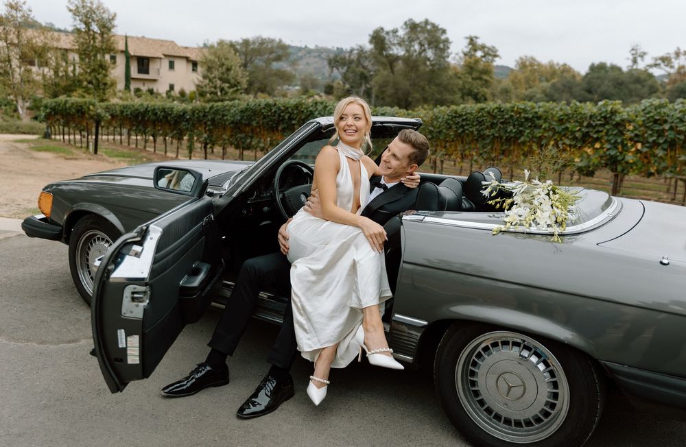 Bride and groom posing with a vintage black Mercedes convertible decorated with white flowers at a vineyard wedding.