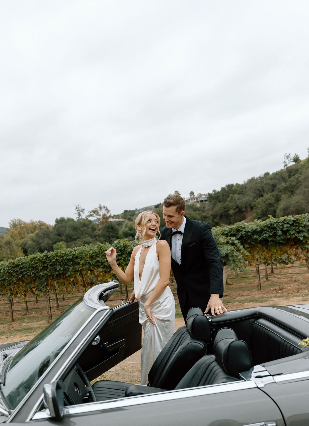 Bride and groom laughing beside a classic convertible car at a vineyard wedding venue.