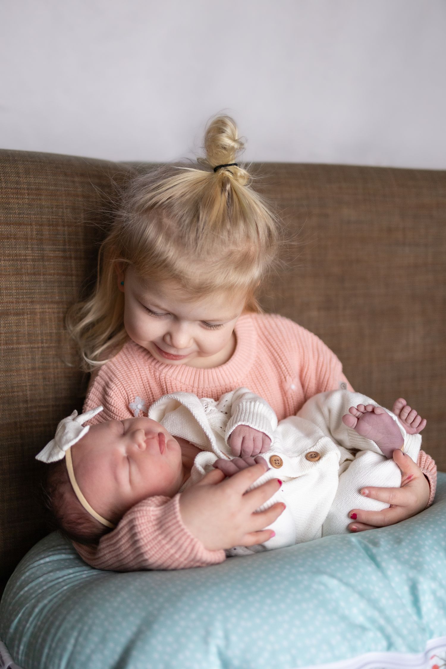 A small child in a pink sweater gently holds and gazes at a sleeping baby in white clothing on a couch.