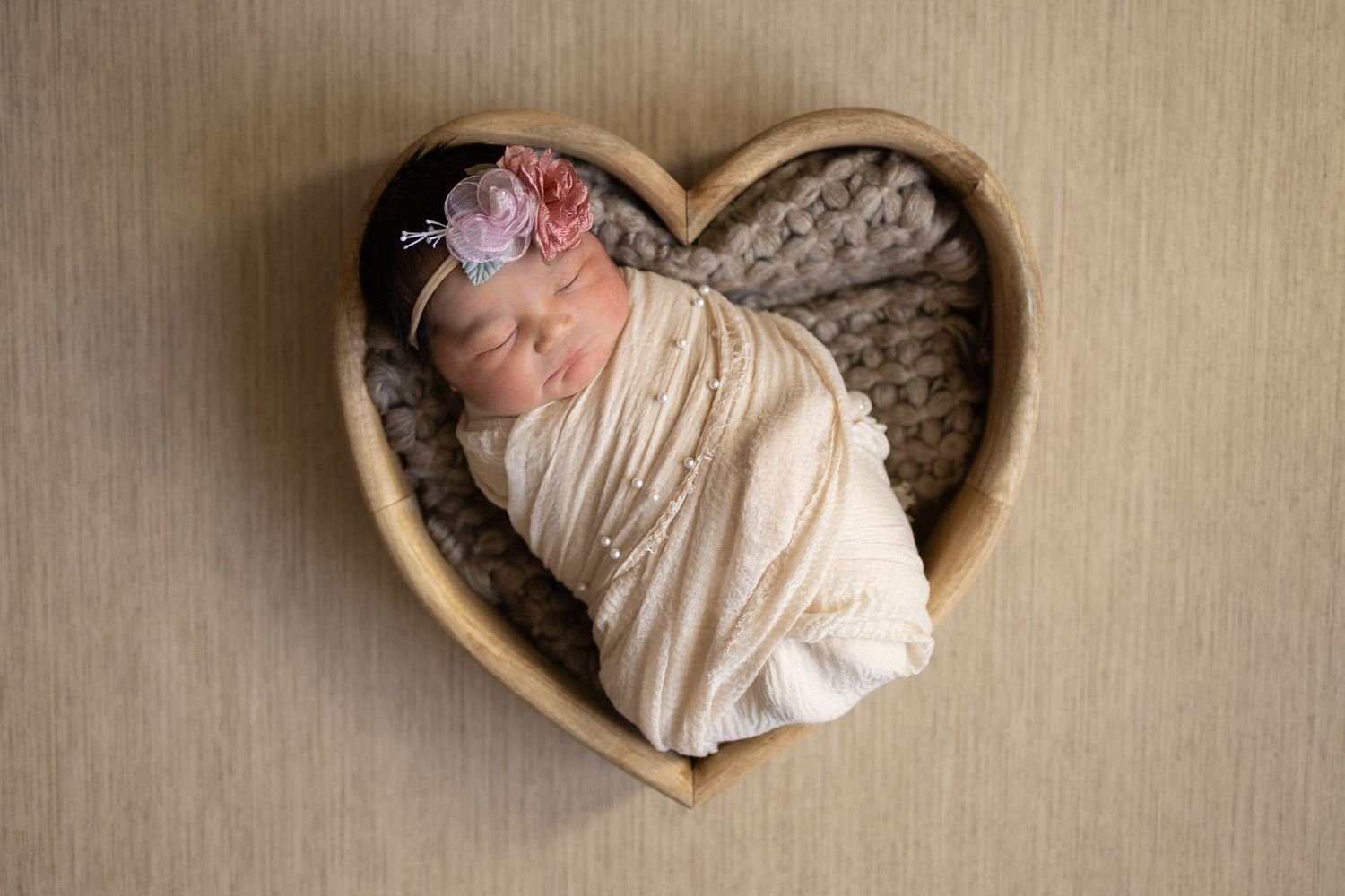 Newborn baby wrapped in cream cloth sleeping peacefully in heart-shaped basket with soft textured blanket.