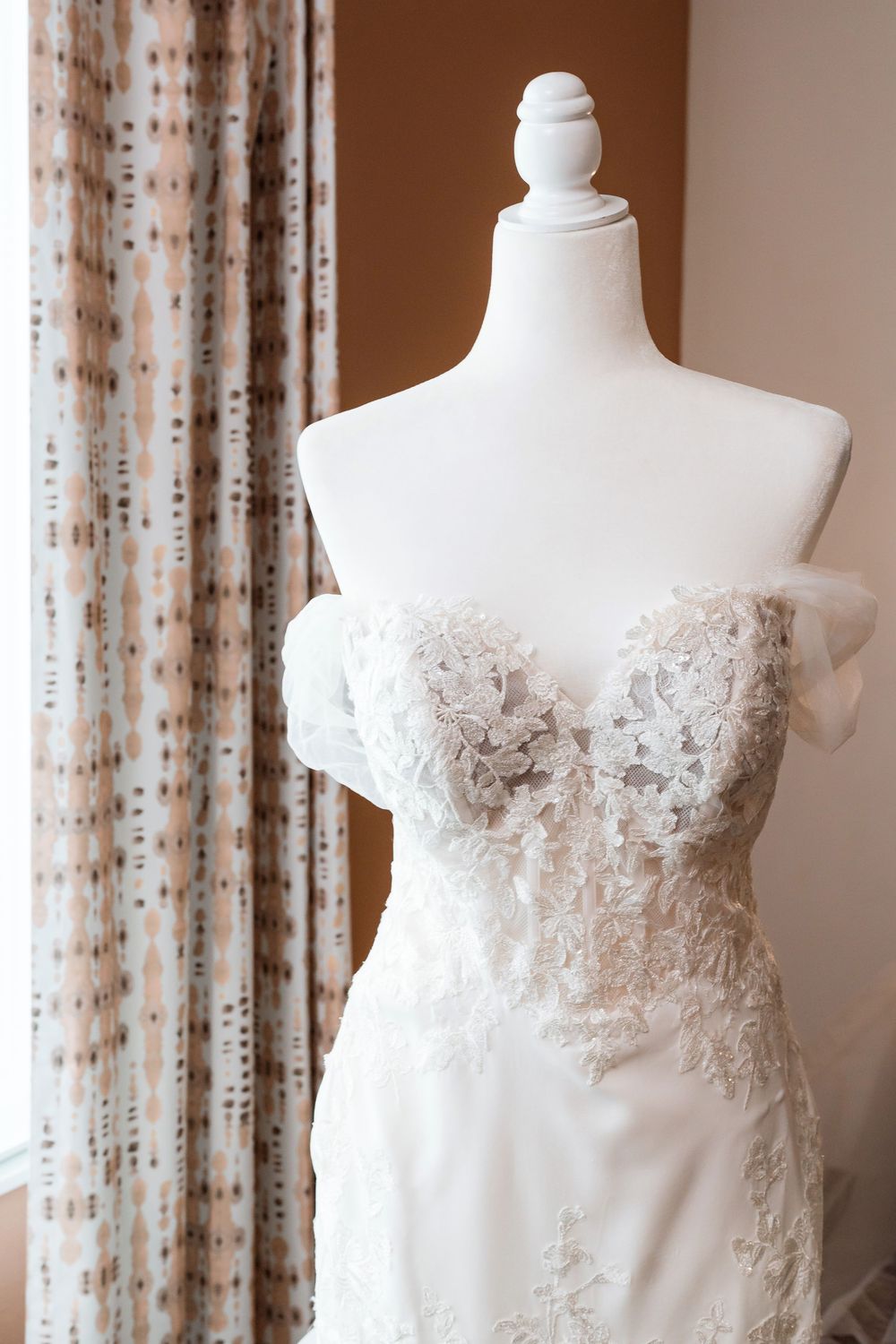 White wedding dress on mannequin display featuring delicate lace bodice detail and flowy fabric near curtained window.