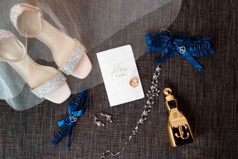 Wedding accessories including blue garters, crystal jewelry, gold charm, ivory heels, and a white card on dark fabric.