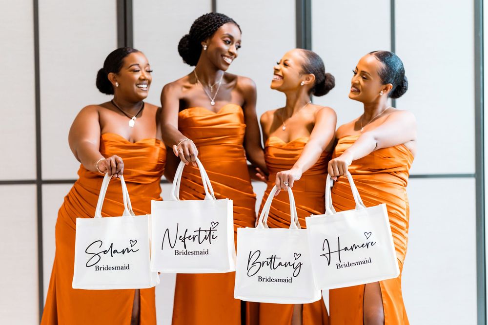 Four bridesmaids in matching orange dresses laugh together while holding personalized white gift bags.