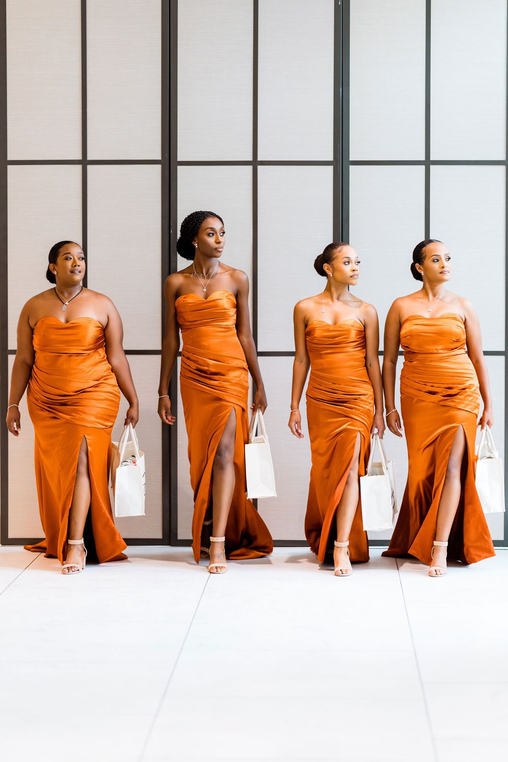 Four graceful dancers in flowing orange gowns pose elegantly against a white modern backdrop with vertical window panels.