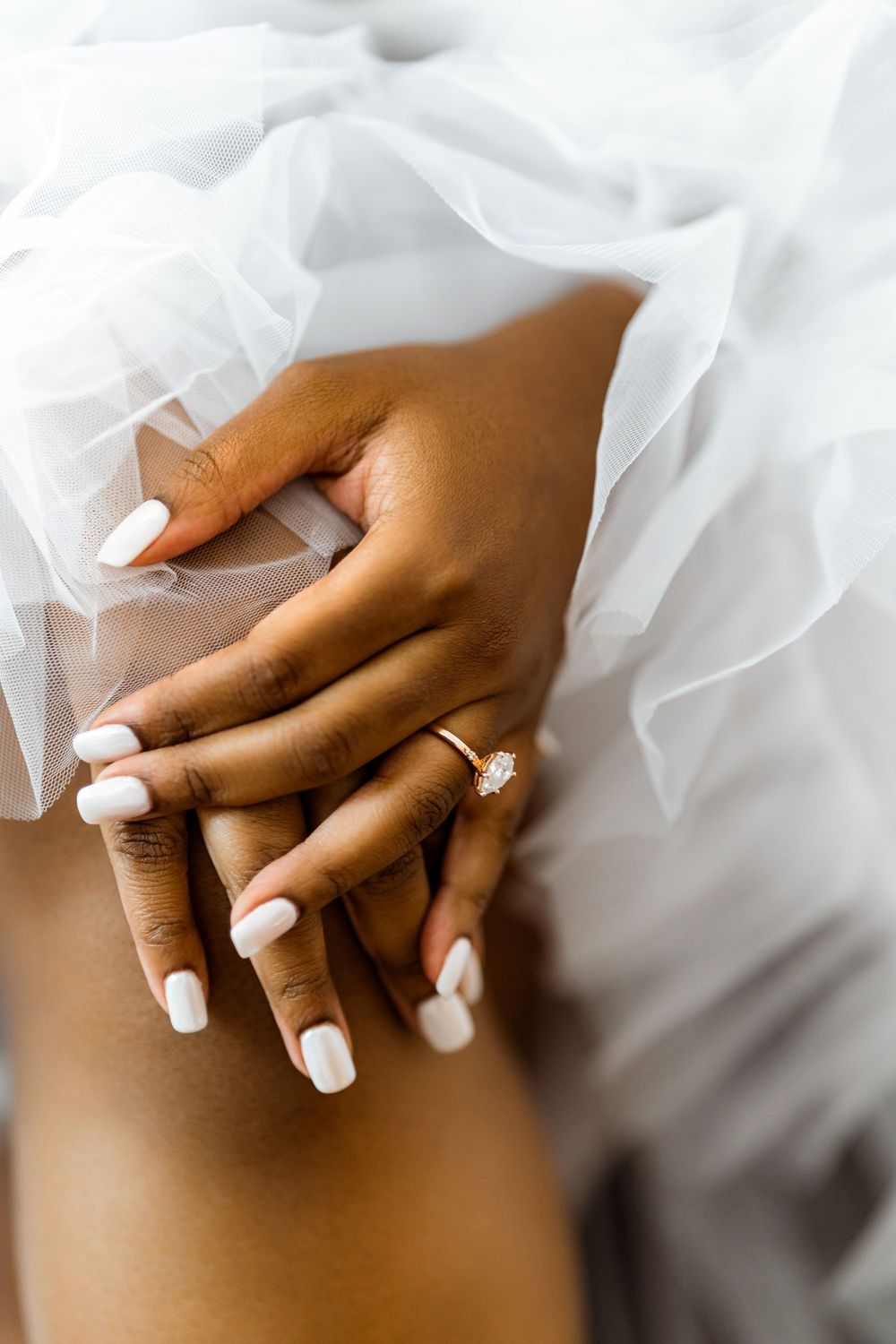Close-up of elegant hands with white manicured nails and diamond engagement ring against white tulle fabric.