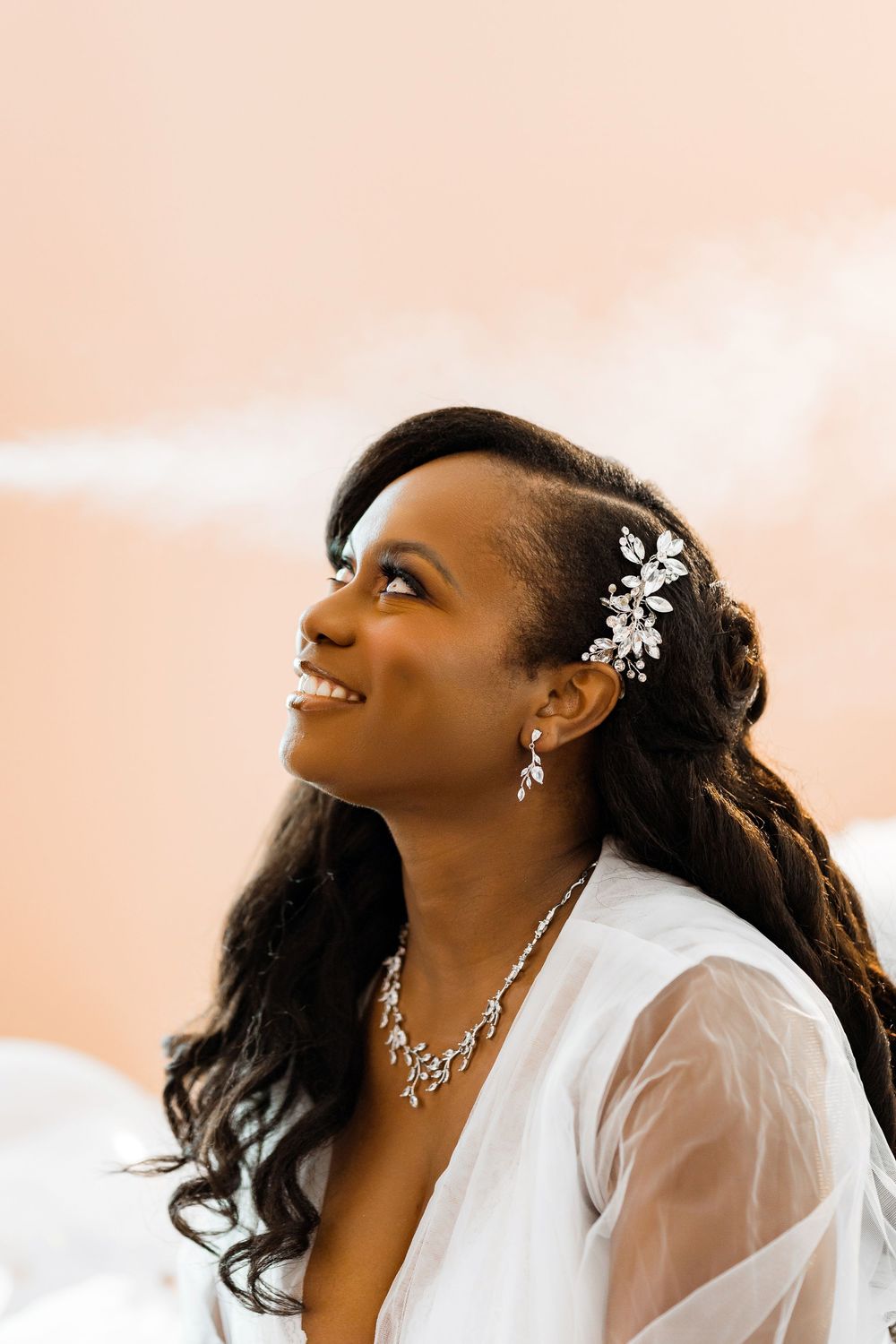 A smiling bride wearing a delicate floral hair accessory and crystal necklace looks up against a peachy sky background.