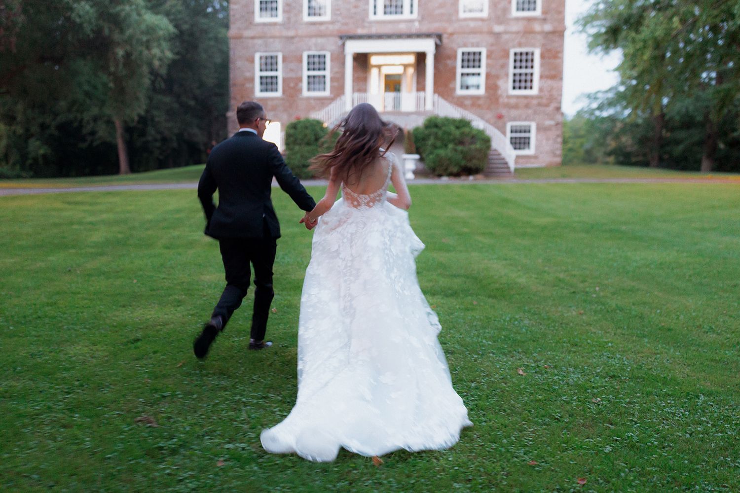 Wedding couple running playfully across a green lawn in front of a brick building.