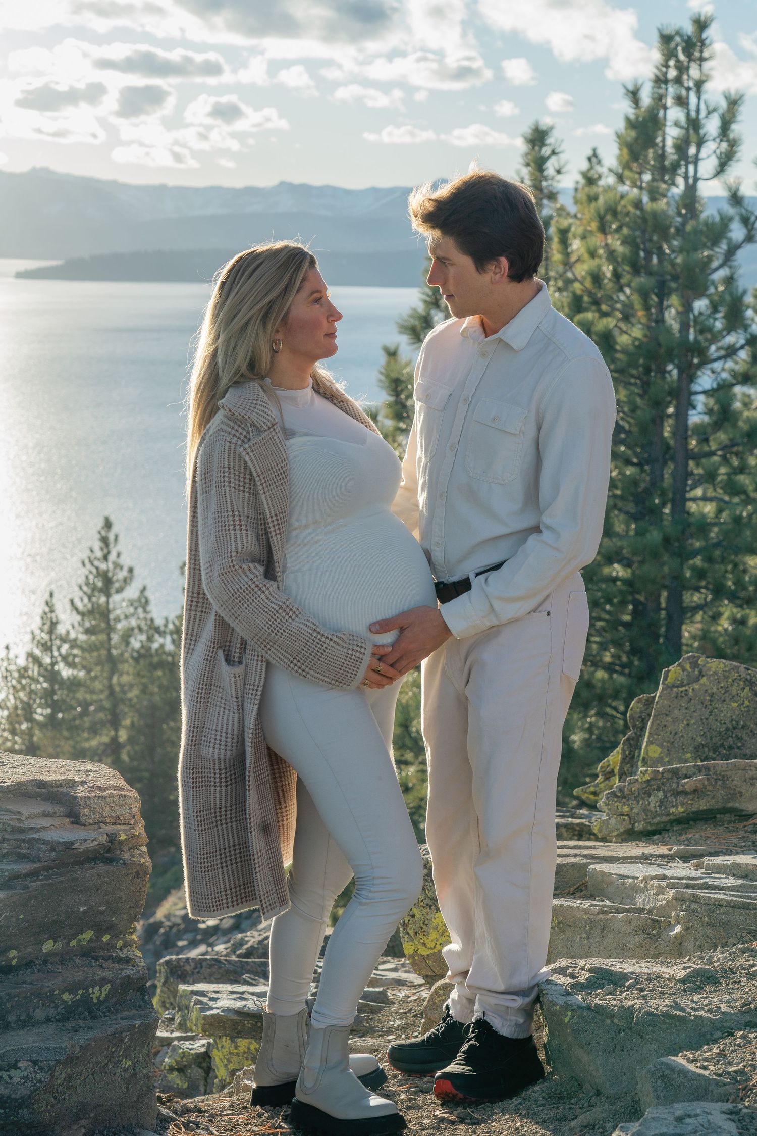 Expectant couple in matching light outfits embrace on mountain overlook with lake and evergreen trees in background.