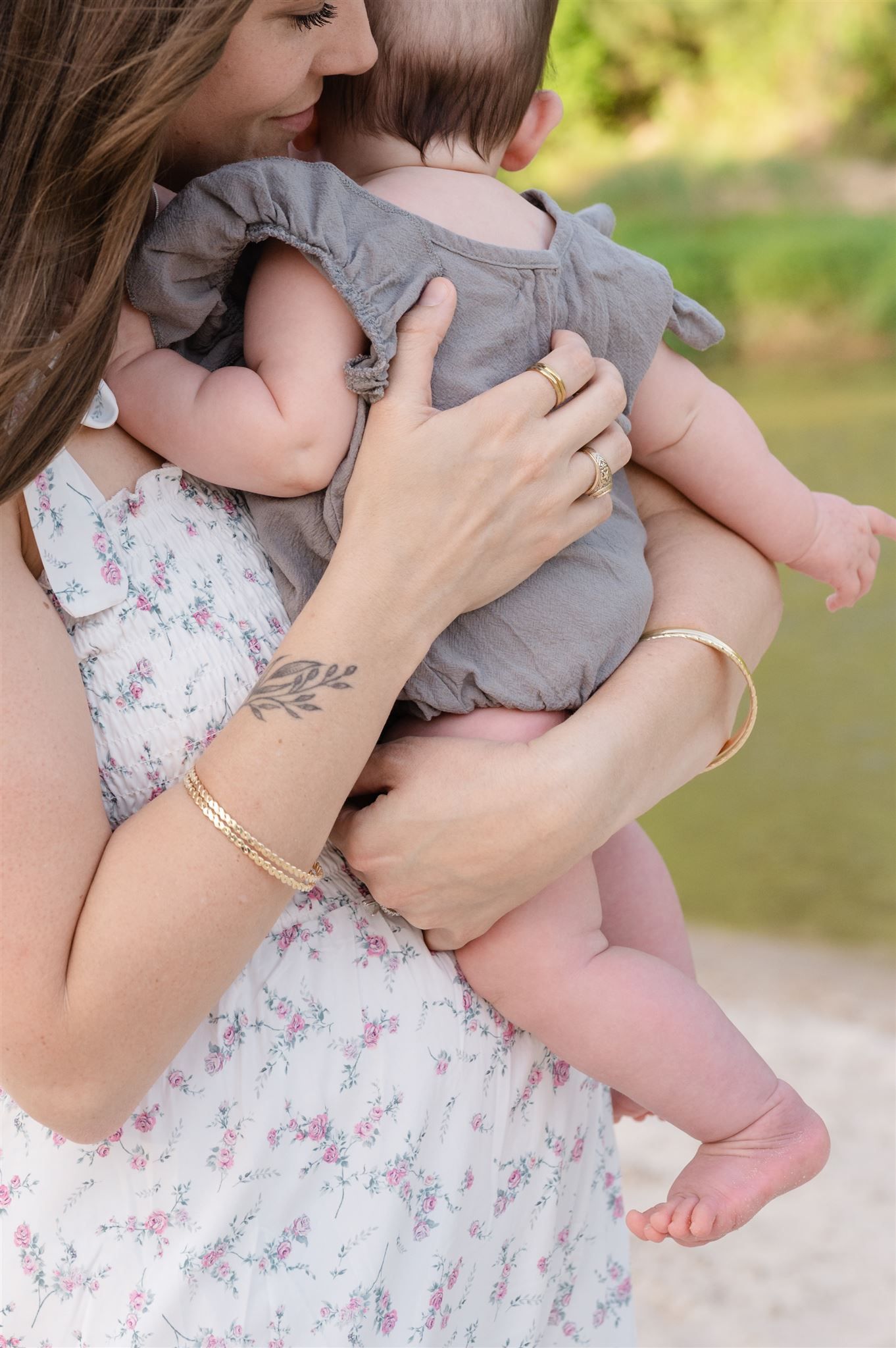 A tender moment captured of someone in a floral dress holding a baby wearing a grey romper outdoors.