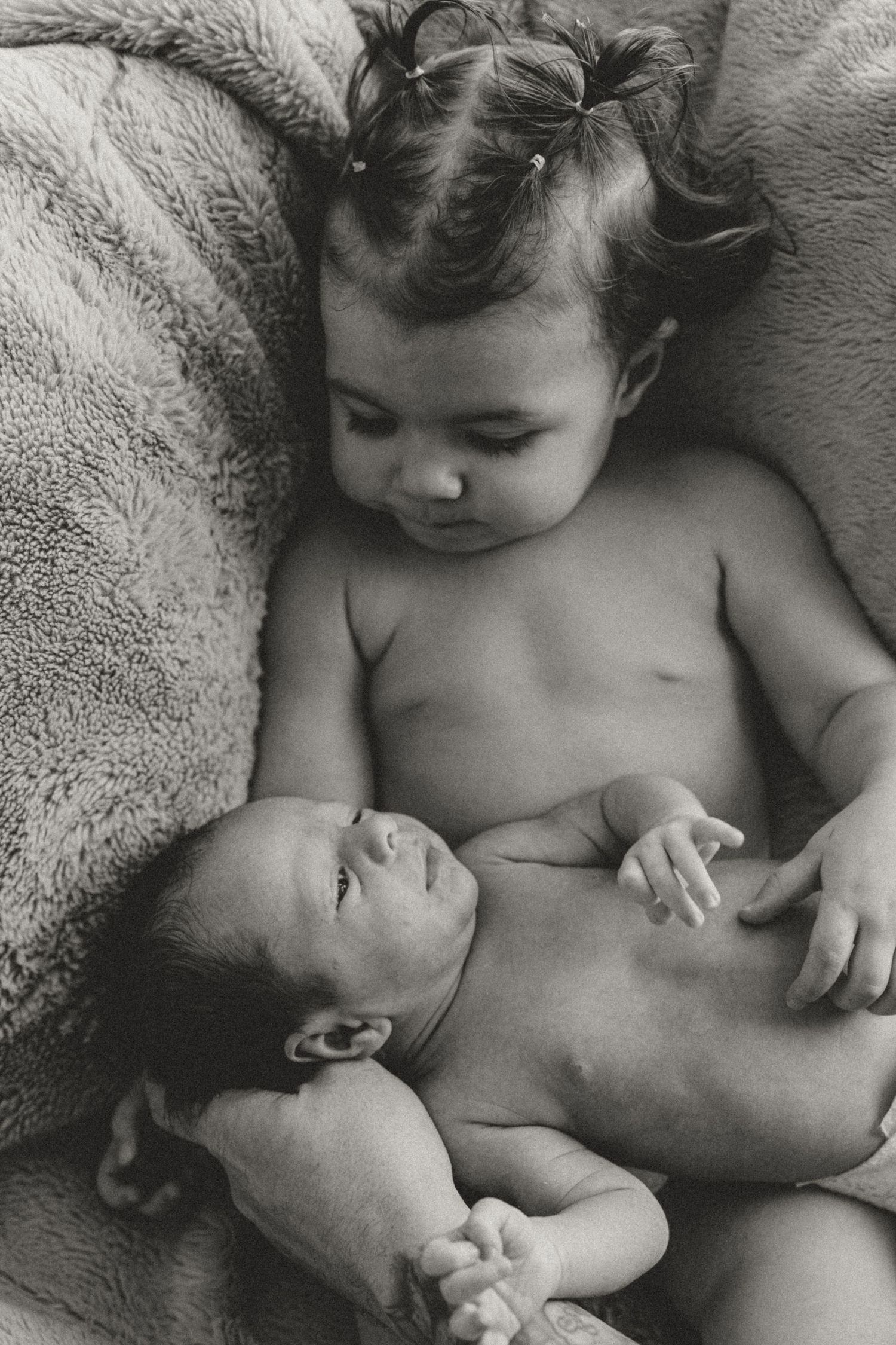 Black and white photograph showing tender sibling moment as baby rests beside toddler on soft blanket.