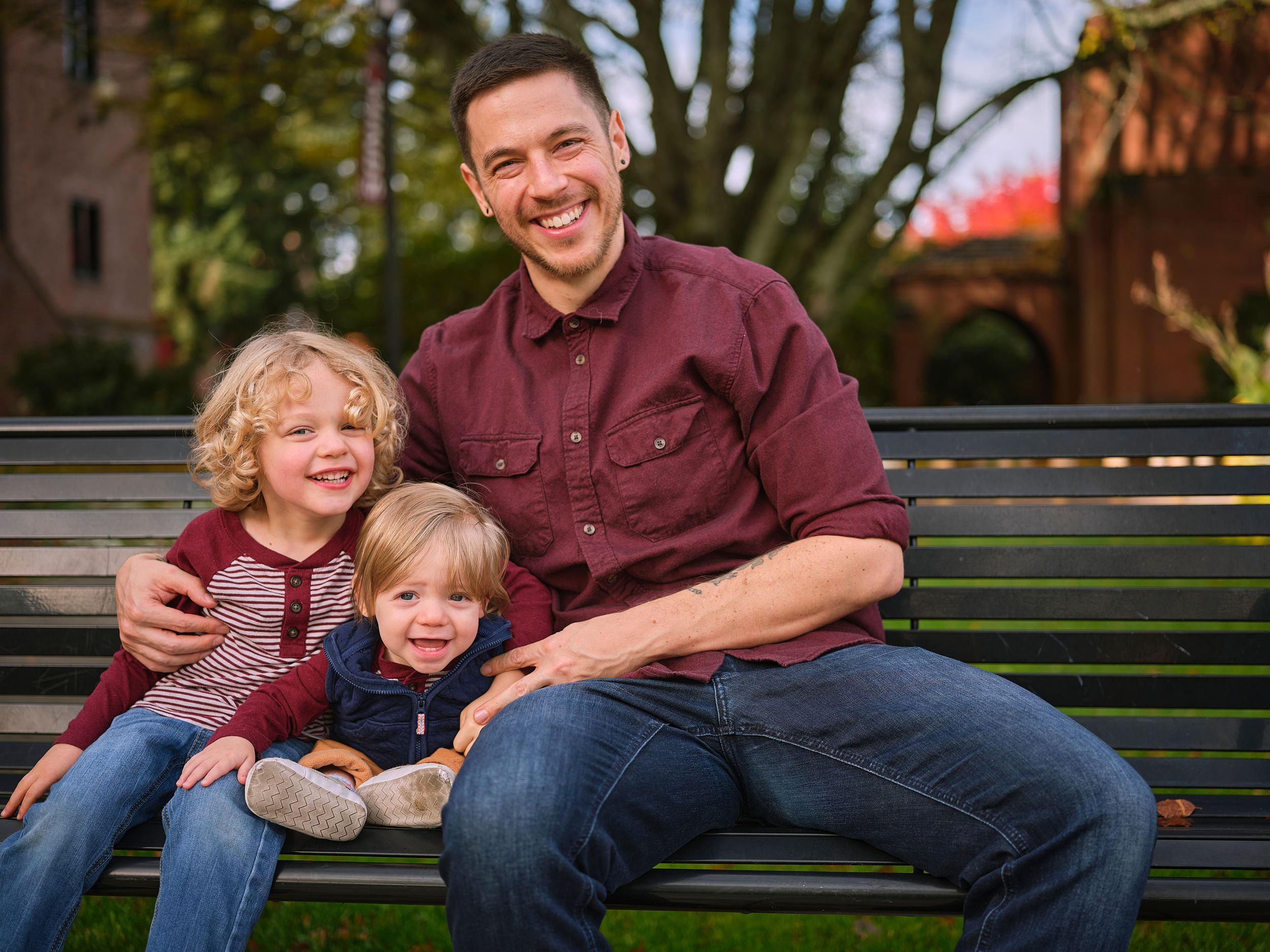 Three people sit together on a wooden park bench surrounded by autumn trees on a sunny afternoon.
