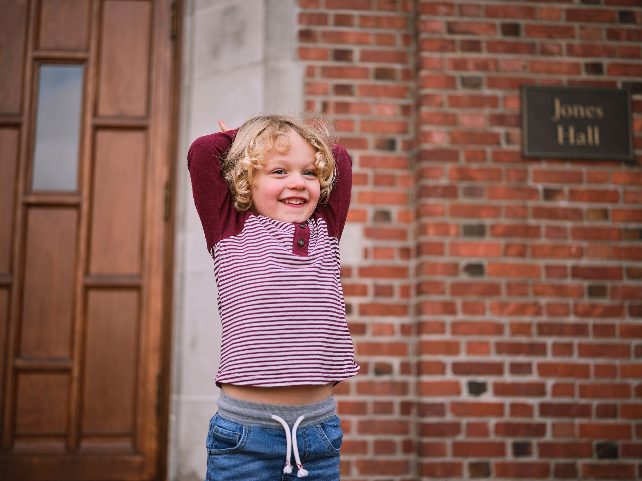 A young child laughs joyfully while playing outside a brick building with wooden doors.