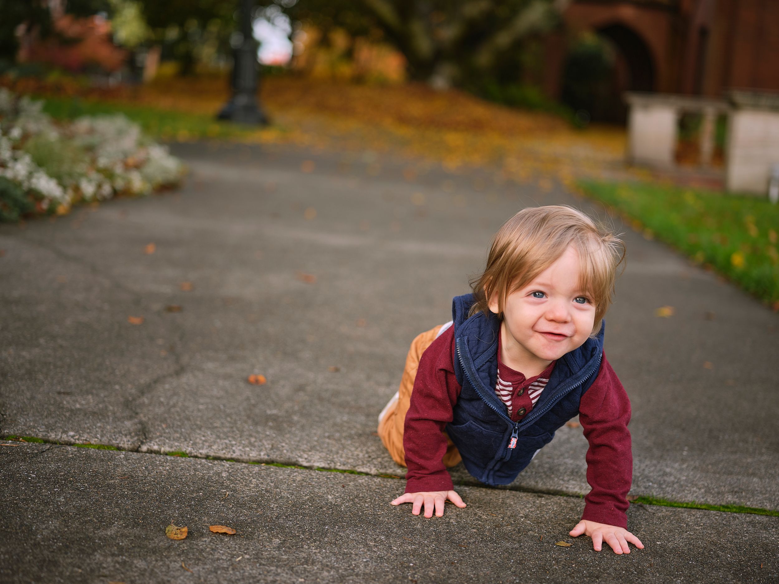 Young child in a navy vest and maroon shirt crawls on a sidewalk with autumn leaves in the background.