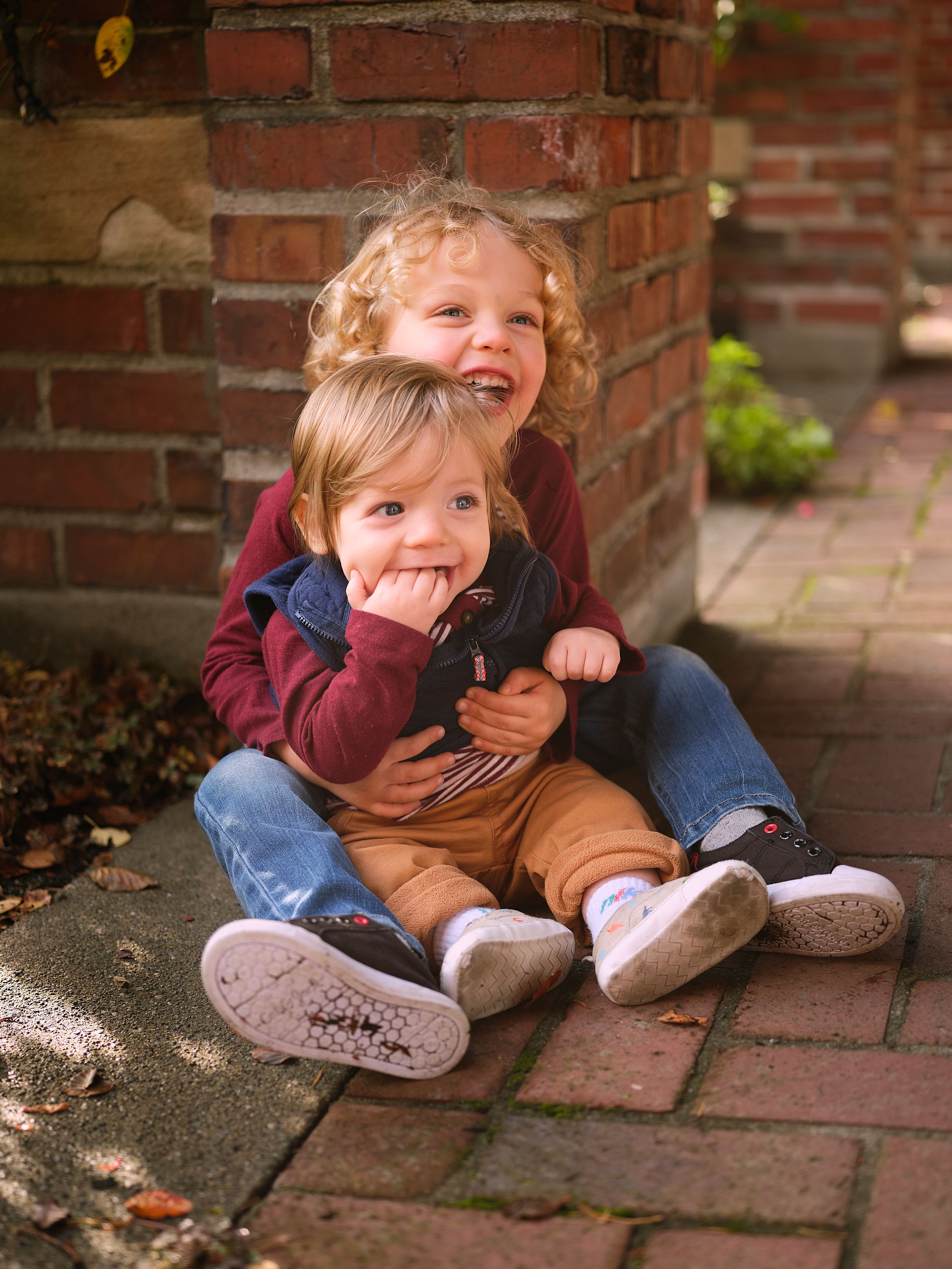 Two kids sitting together on brick pavers and laughing while wearing casual fall clothing.