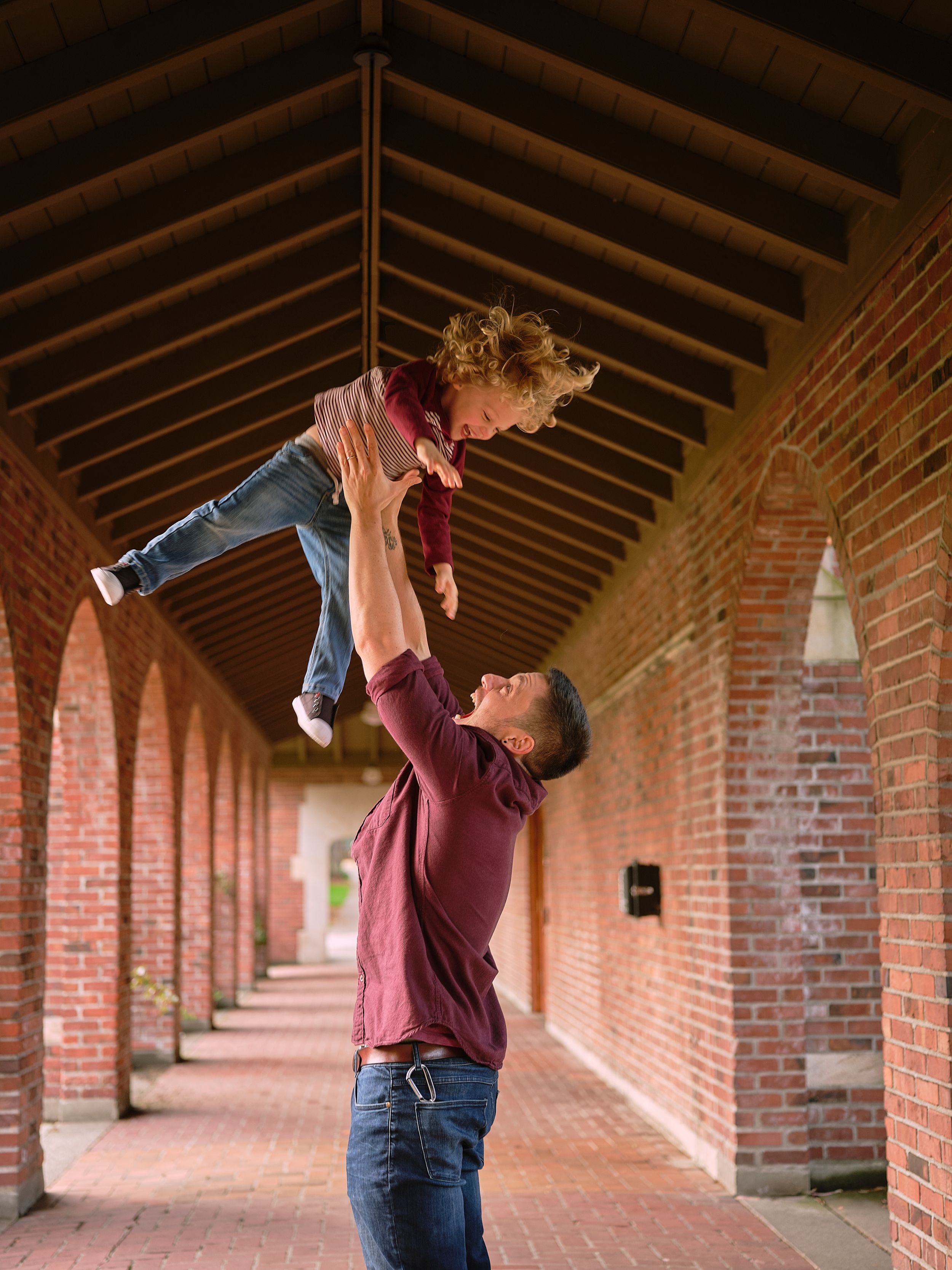 Parent lifts small child playfully in a covered brick archway corridor with exposed wooden beam ceiling.