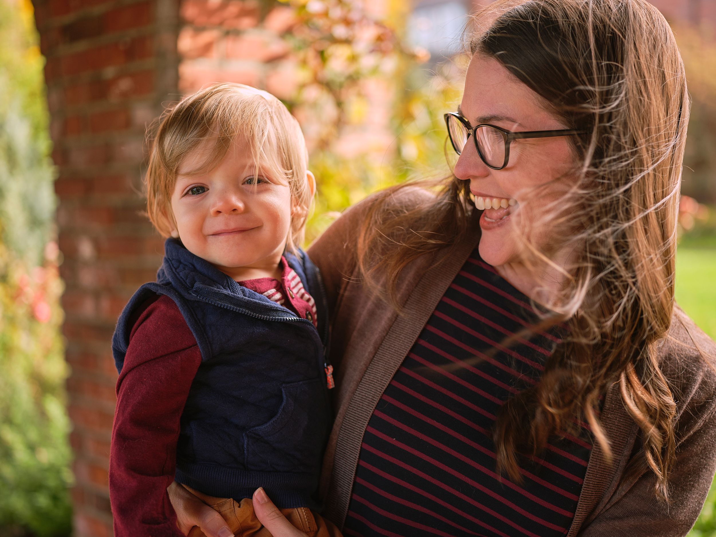 A woman in glasses and striped shirt shares a joyful outdoor moment with a small child in a navy vest on an autumn day.