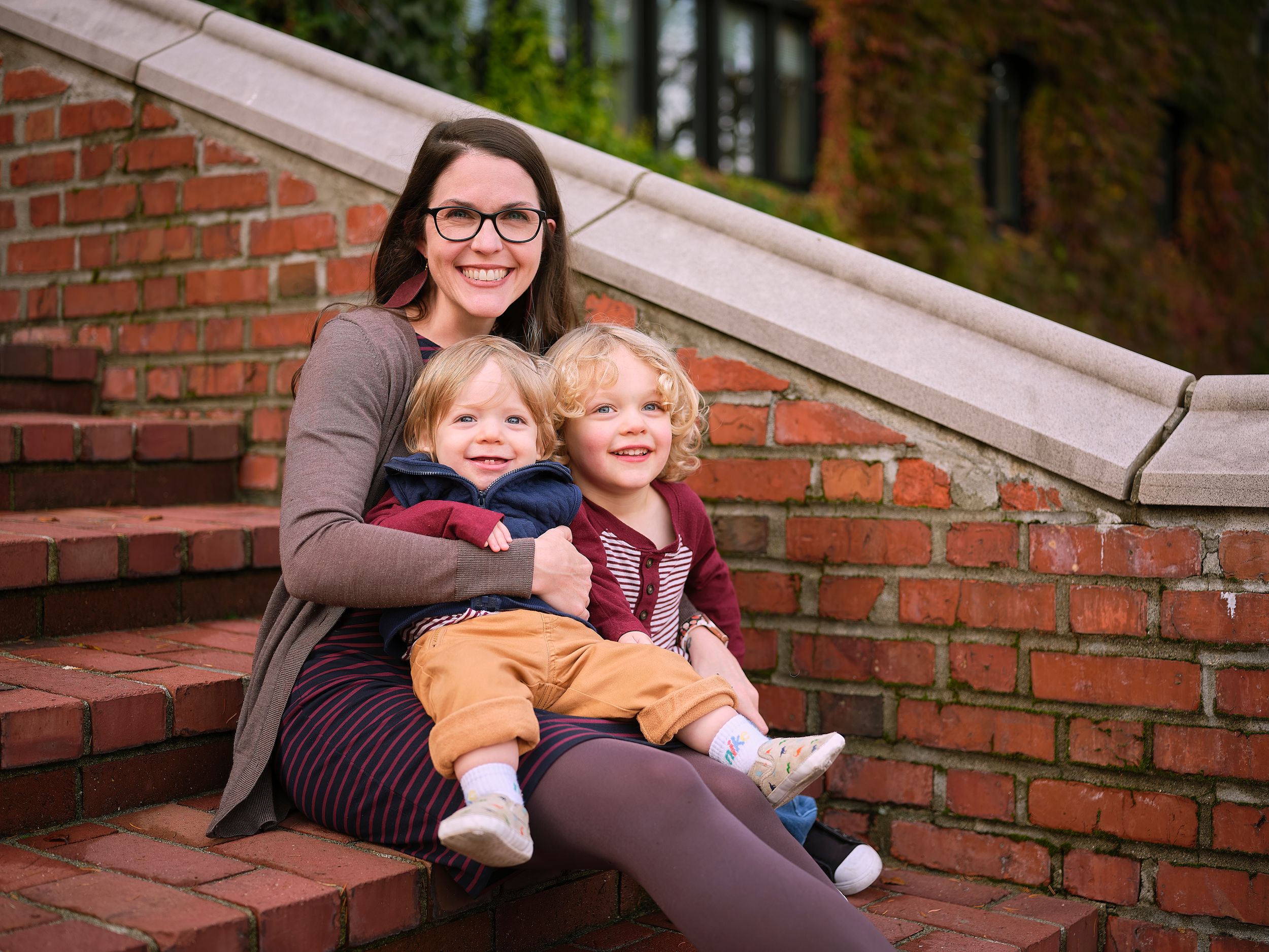 A parent and two young children sit together on outdoor brick steps on an autumn day.