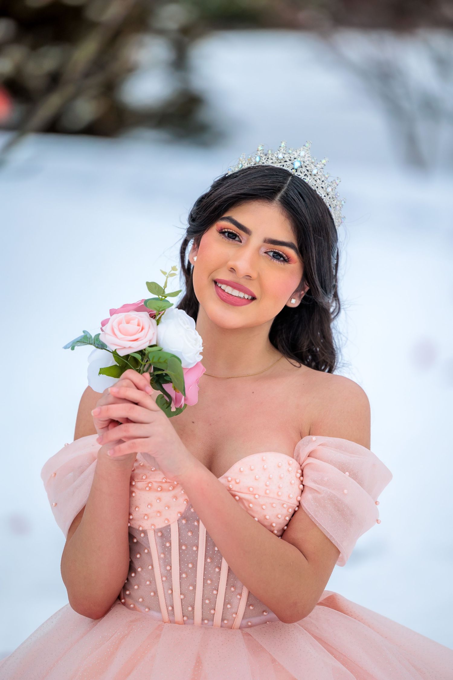 Young woman in pink quinceañera ball gown wearing a tiara and holding roses in a snowy outdoor setting.