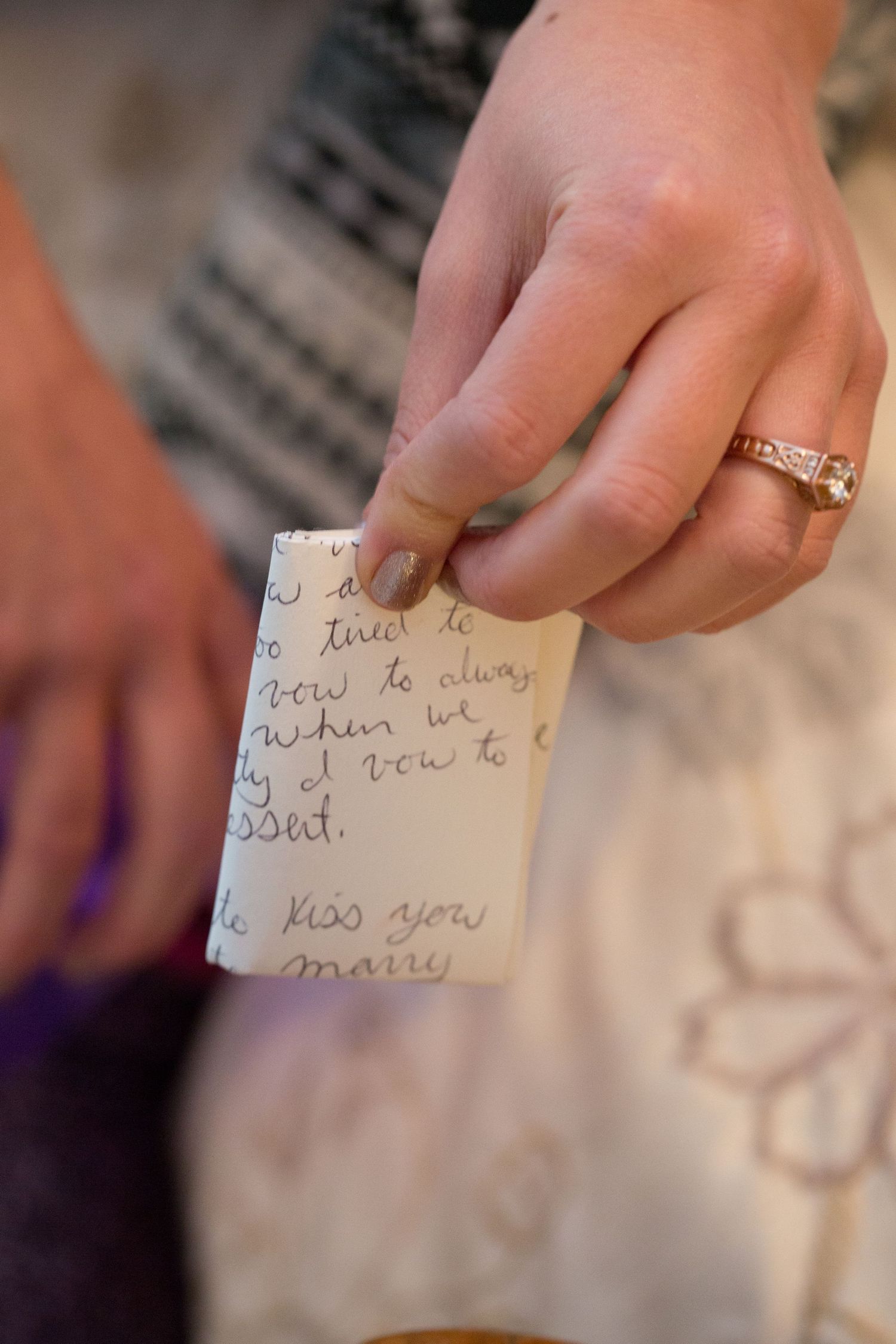A hand wearing an engagement ring holds a small handwritten note with text visible on white textured fabric.