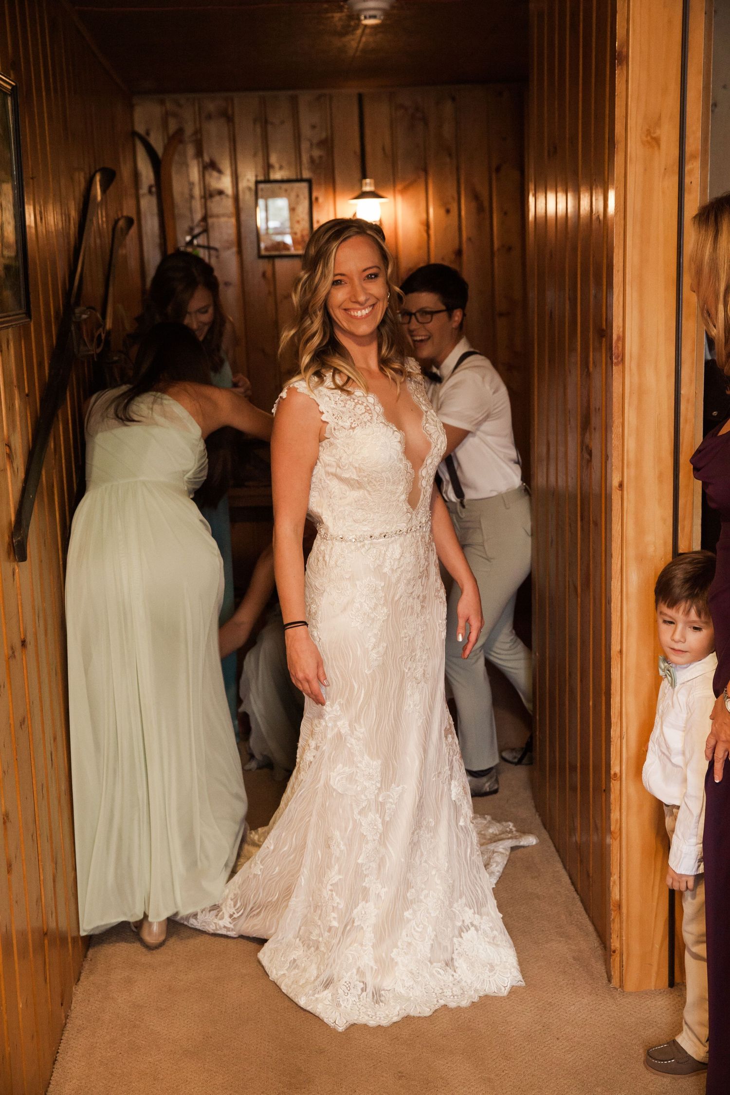 A person in a stunning white lace wedding dress smiles while standing in a wood-paneled room with others visible nearby.