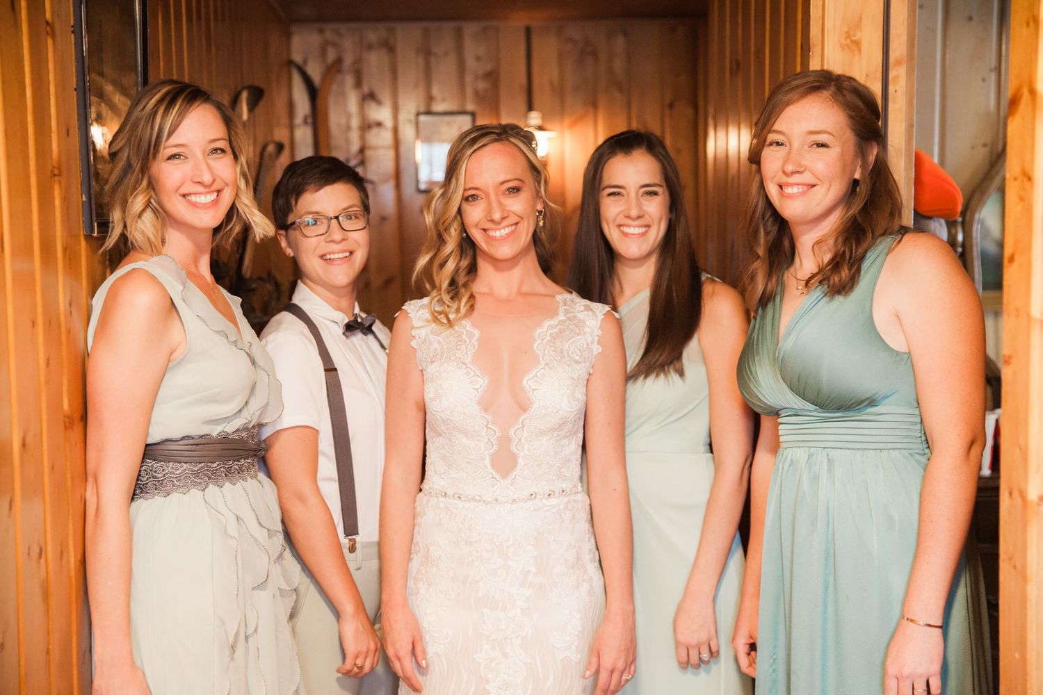 Bridesmaids in sage green and ivory dresses pose with bride in lace gown inside wood-paneled room with warm lighting.
