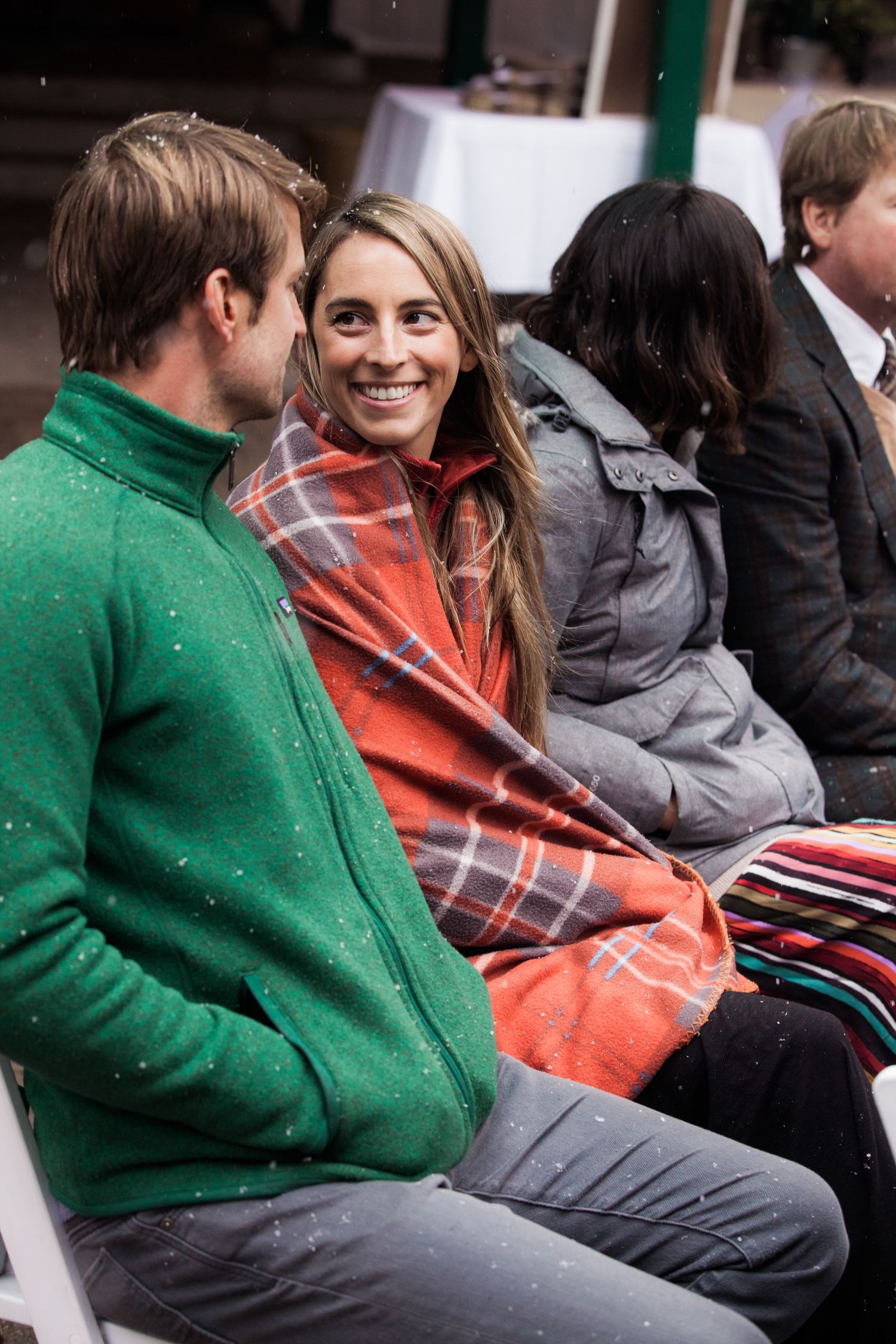 A couple sits together outdoors, wearing winter attire with the person in a green sweater and the other in a plaid scarf.
