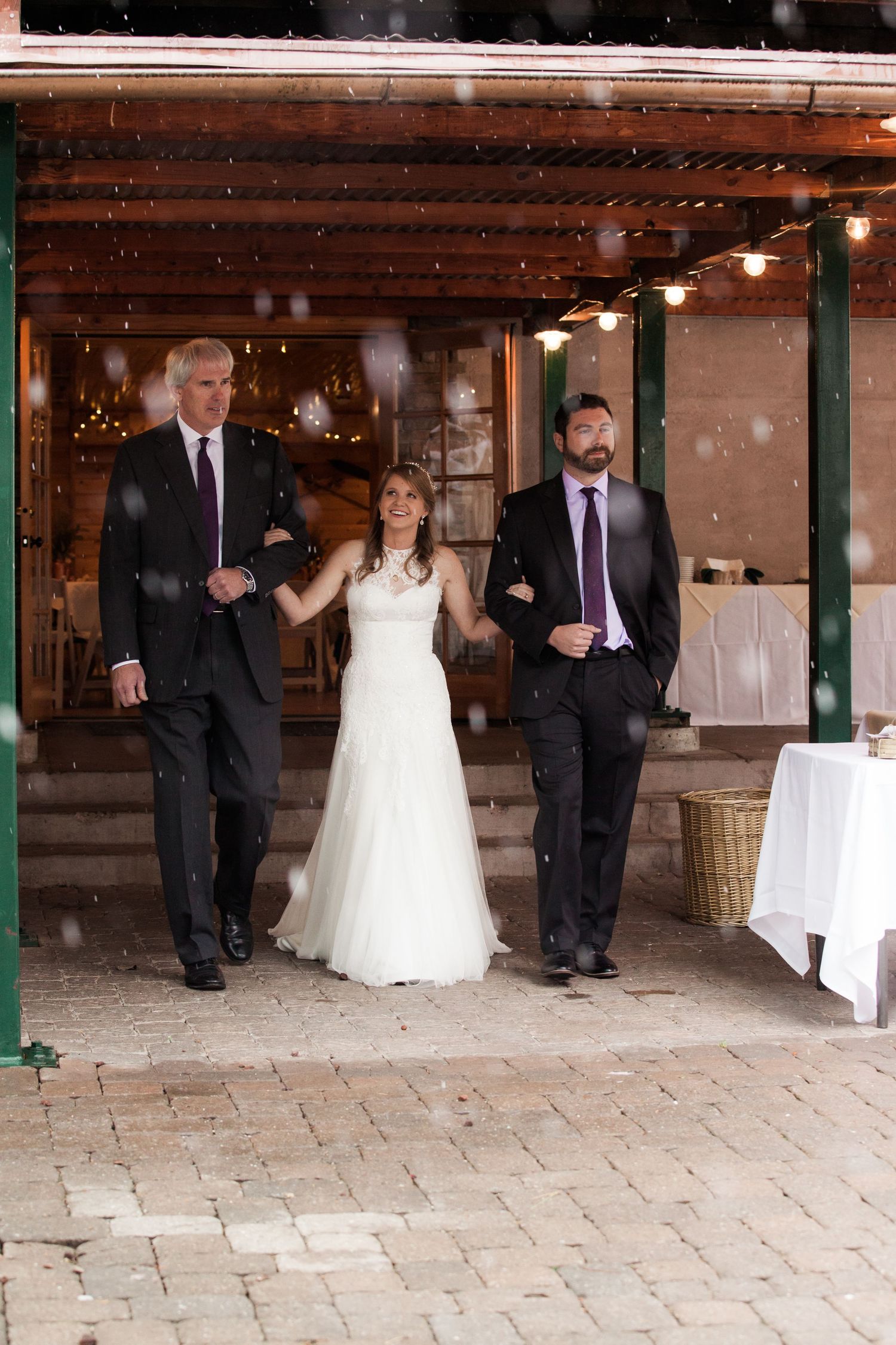 Wedding party stands under covered porch with string lights during light snowfall at outdoor winter ceremony.