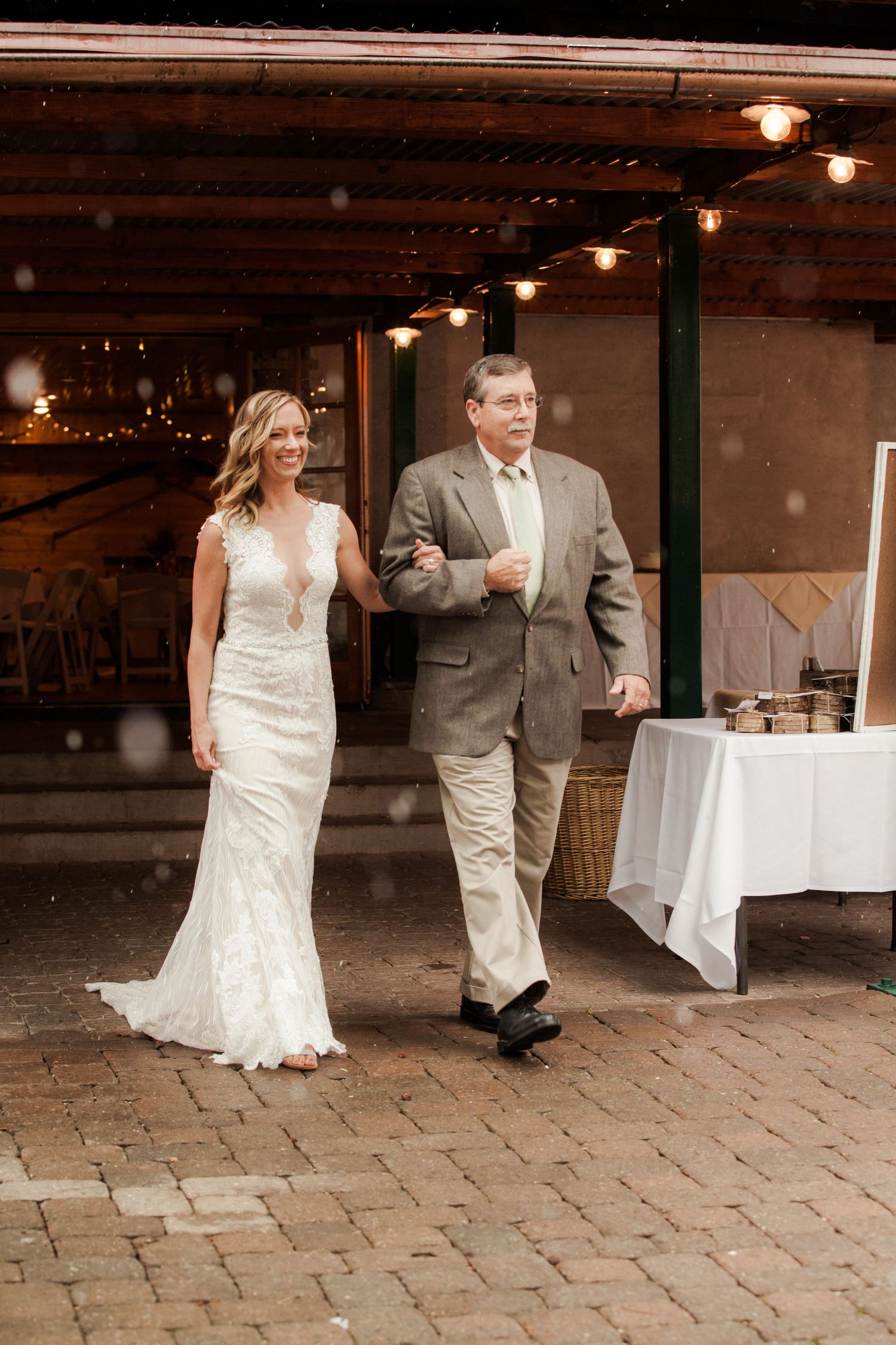 A bride in a fitted lace gown walks alongside a man in a gray suit under string lights on a brick patio.