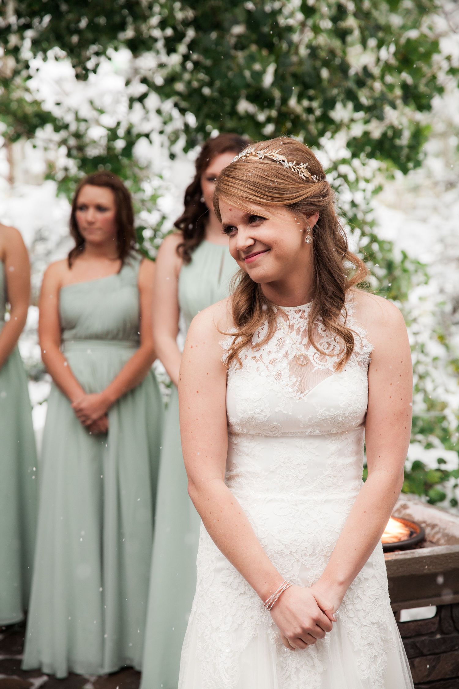 Bride in white lace dress smiles with bridesmaids in sage green dresses standing behind at outdoor wedding ceremony.