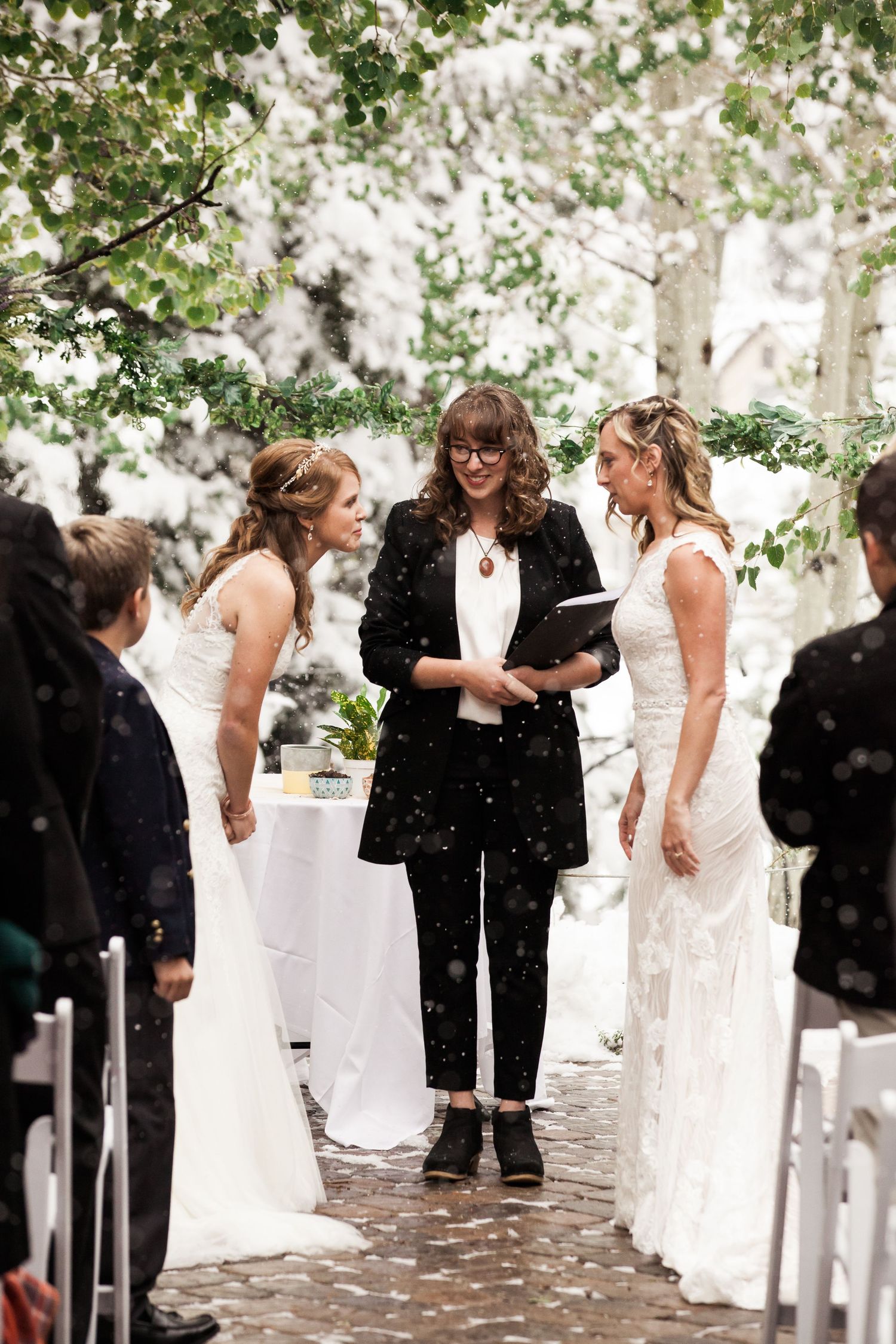 An outdoor wedding ceremony takes place in a garden setting with white chairs and green foliage overhead.