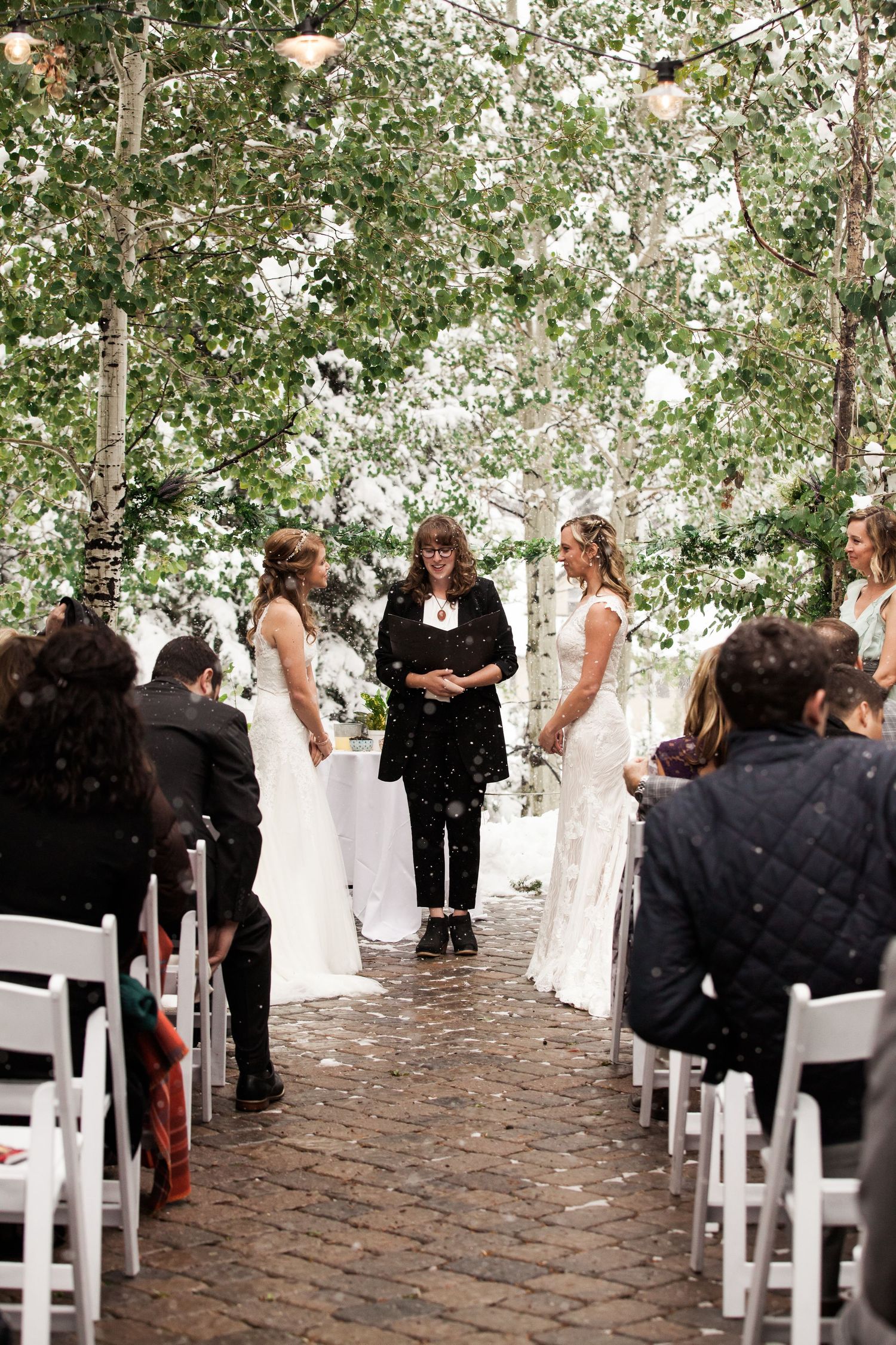 Two people in white dresses face an officiant during an outdoor wedding ceremony on a cobblestone path under string lights.