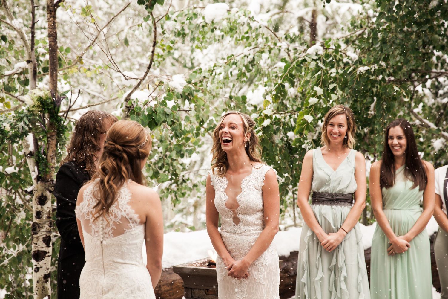 A joyful wedding party shares a candid moment in an outdoor garden setting with bridesmaids in sage dresses.