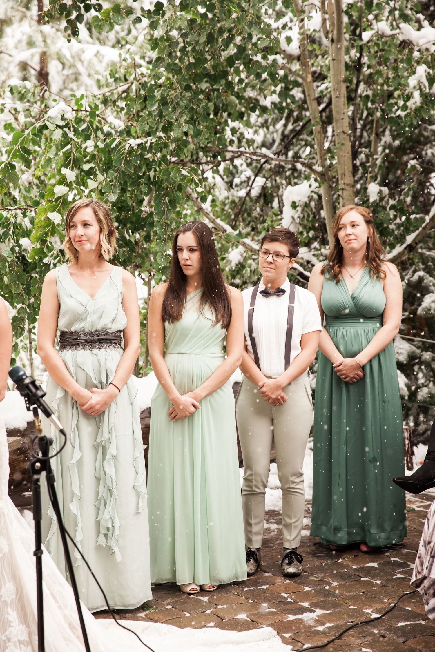 Wedding party members stand together in mint and sage colored dresses with a snowy winter backdrop during outdoor ceremony.