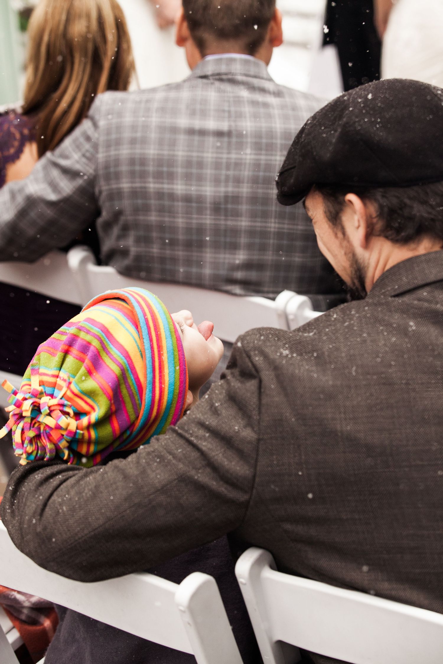 A person in a winter hat with colorful stripes and pom pom sits in a white chair during a snowy outdoor event.