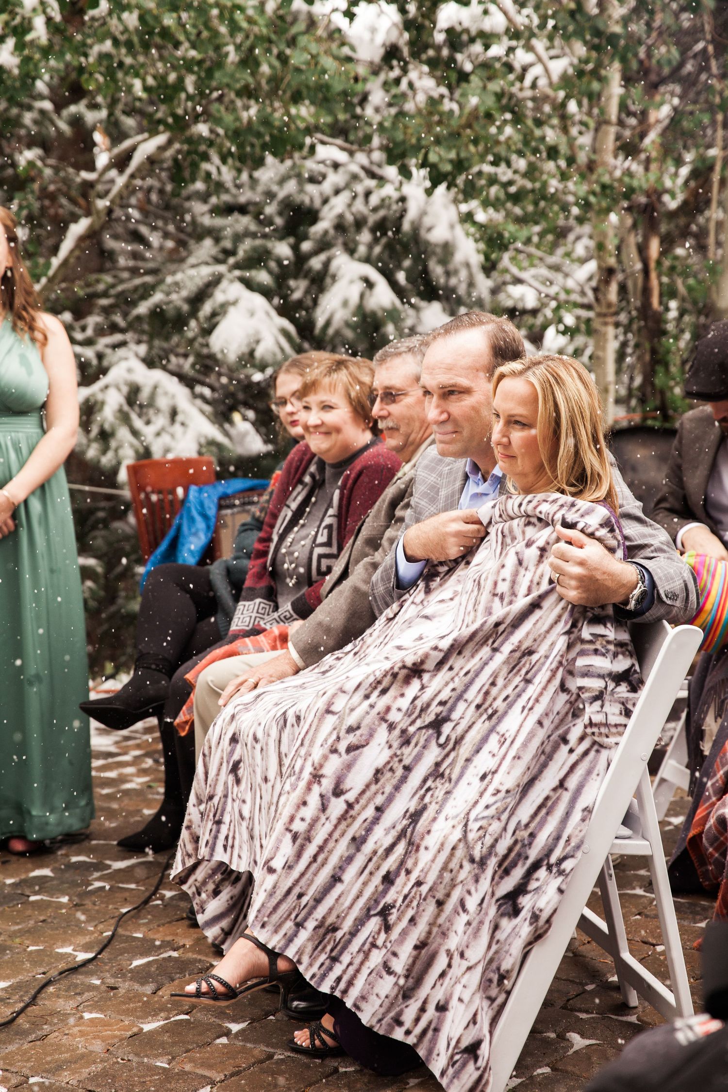 Guests sit bundled in blankets at an outdoor winter gathering with snow-covered trees in the background.