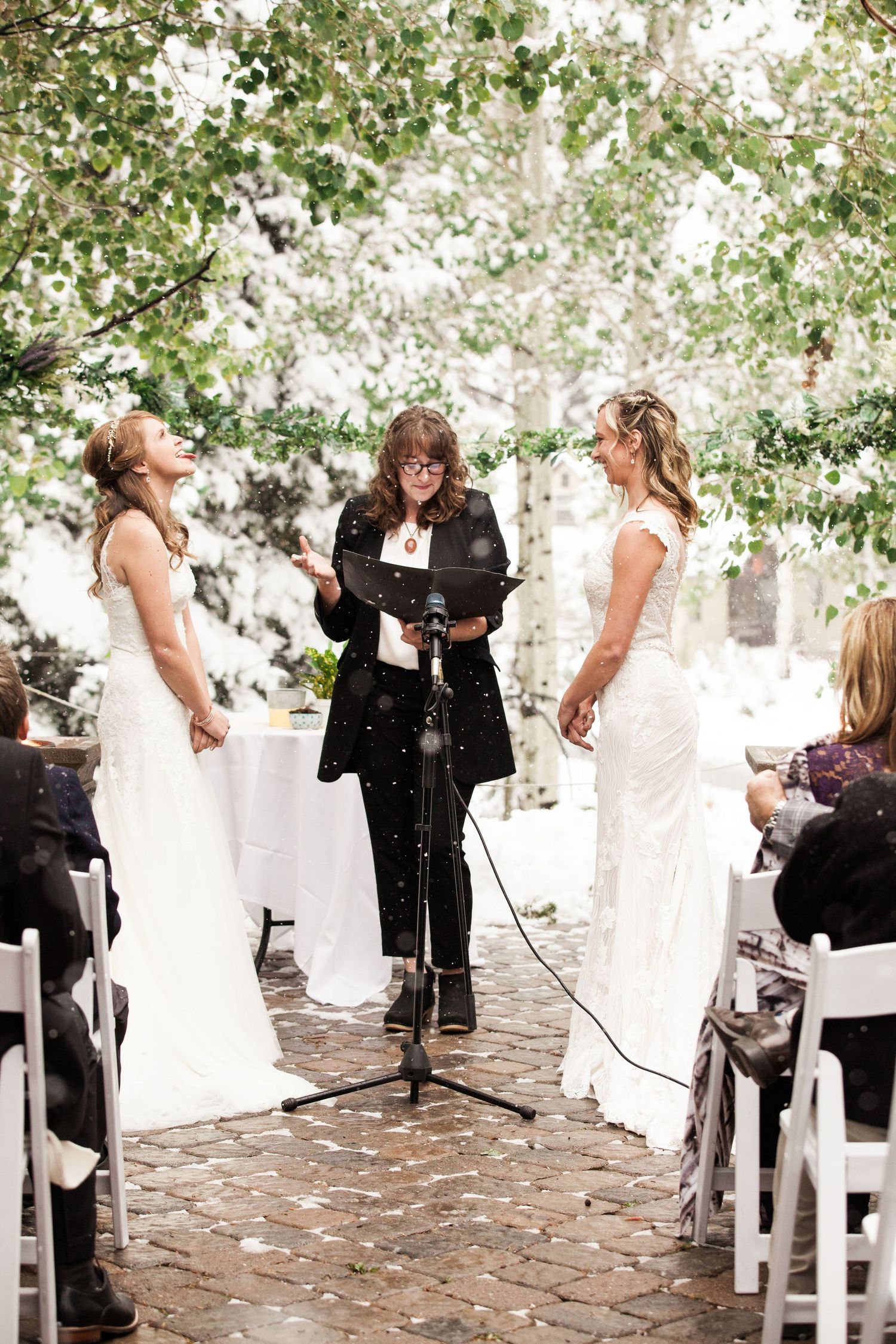 Two brides share a moment during their outdoor ceremony while an officiant in black reads from a podium.