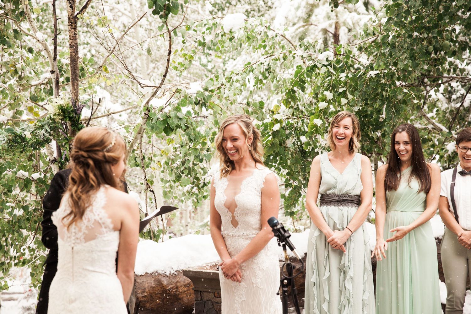 A group in light-colored dresses stands together outdoors in a natural setting surrounded by trees and sunlight.