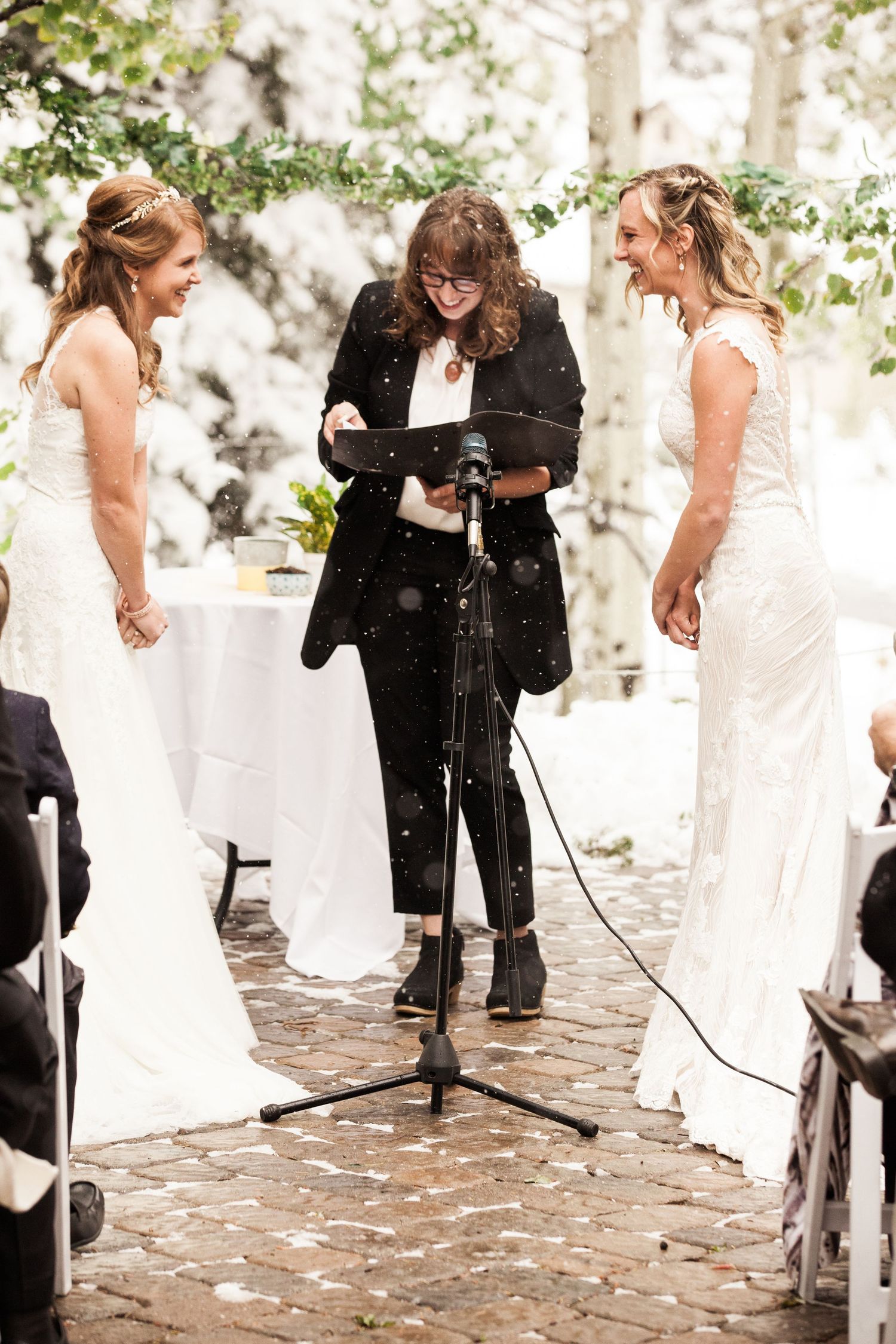 Two brides in white dresses stand with an officiant in black during an outdoor wedding ceremony on a brick patio.
