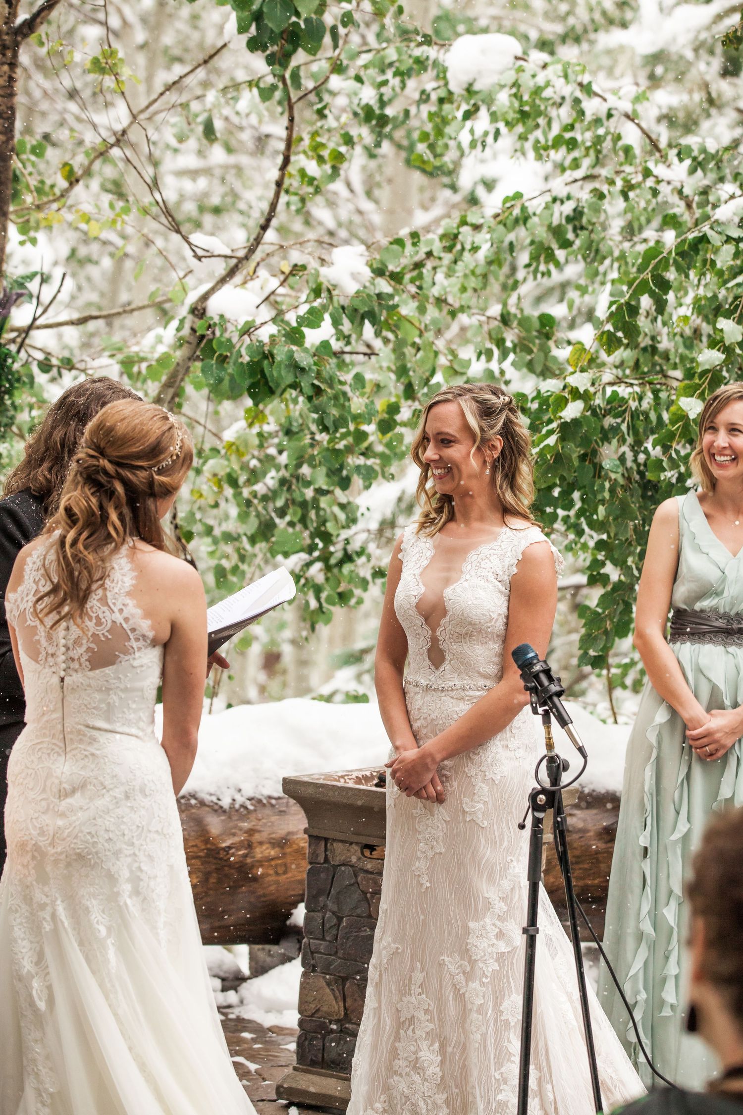 A wedding ceremony takes place outdoors with delicate lace gowns against a backdrop of green foliage.