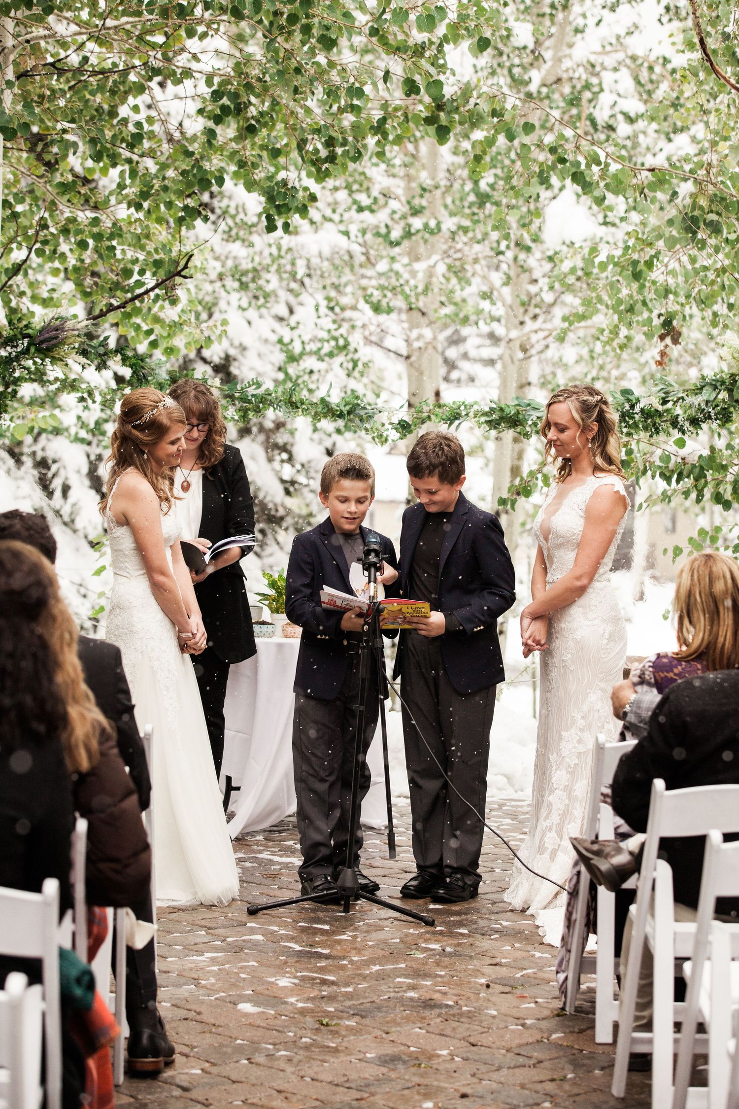 An outdoor wedding ceremony with guests seated on white chairs while young participants read from papers.