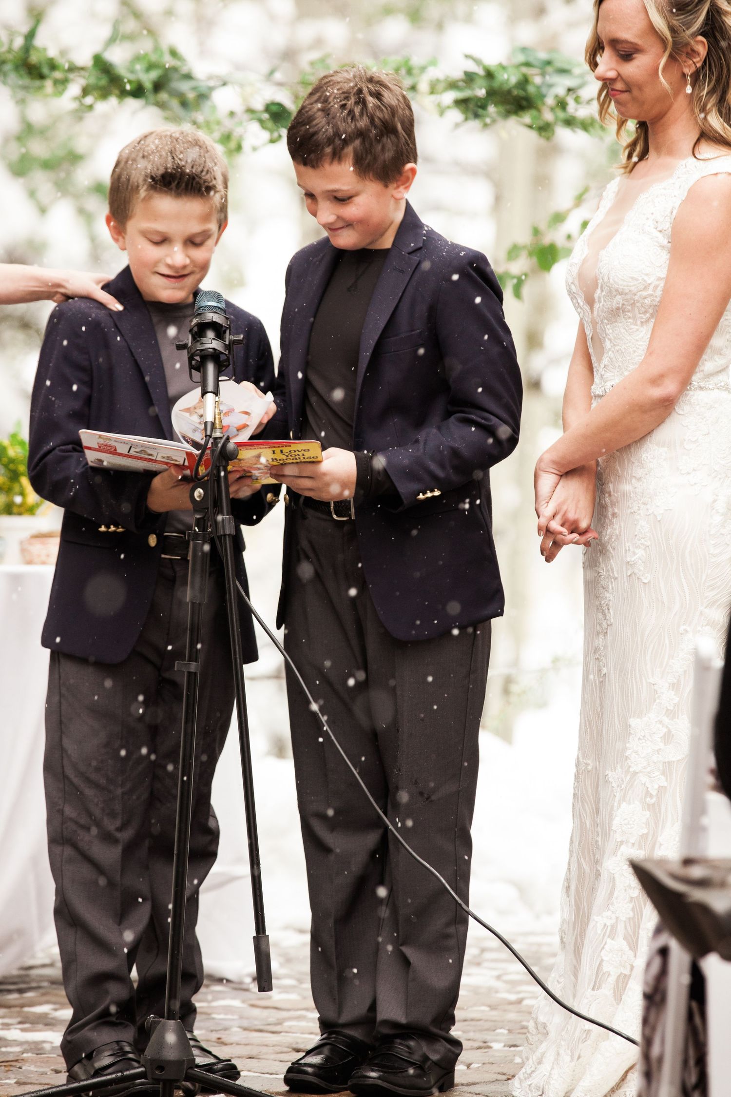 Two young members of the wedding party read from a shared document during an outdoor ceremony with falling snow.