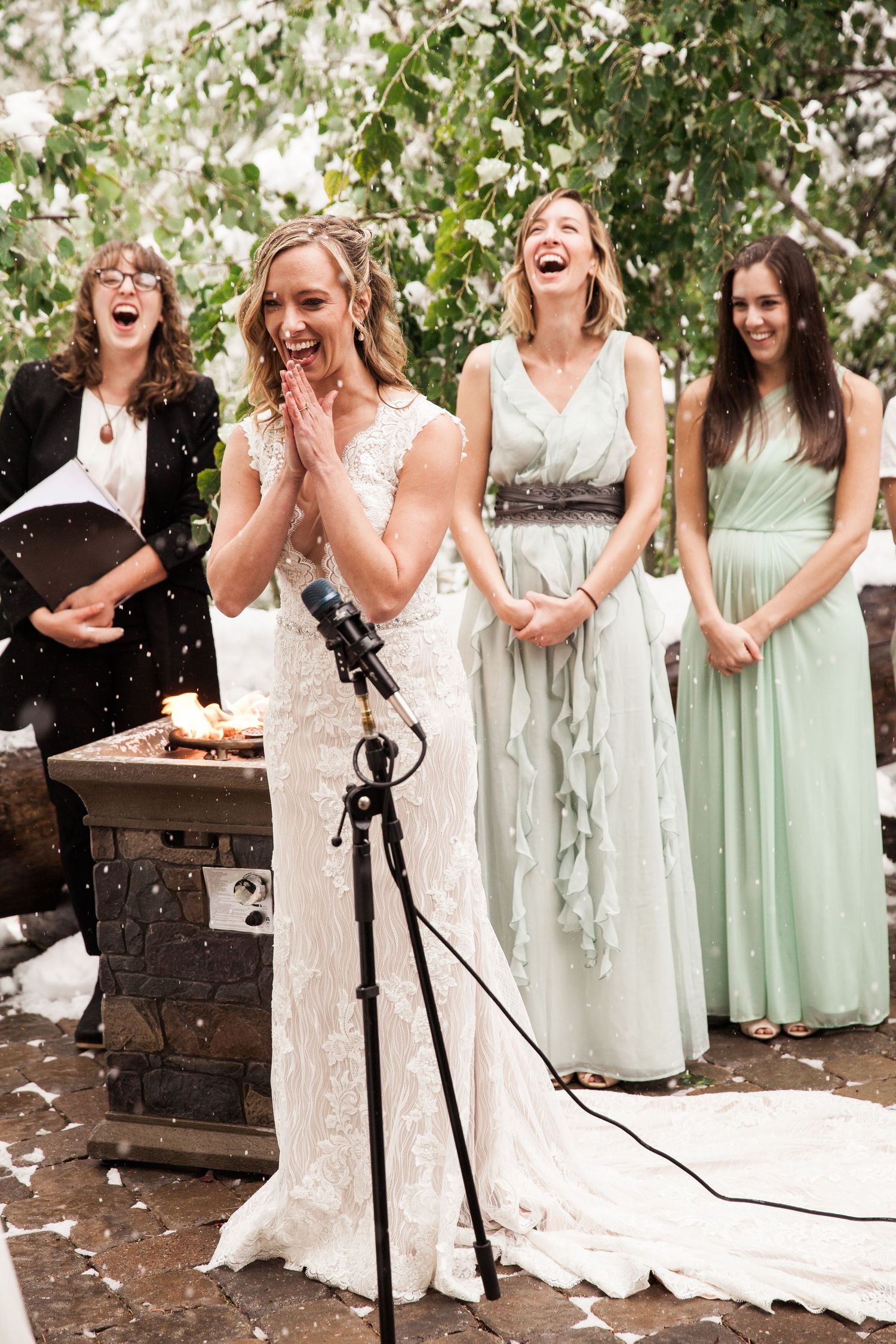 A joyful wedding celebration with people in elegant dresses laughing together outdoors under leafy trees.