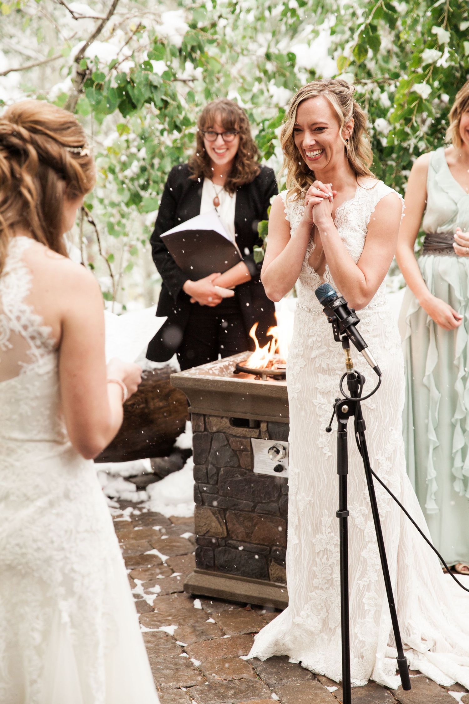 Two people in white wedding dresses share a joyful moment during an outdoor ceremony with snow falling.