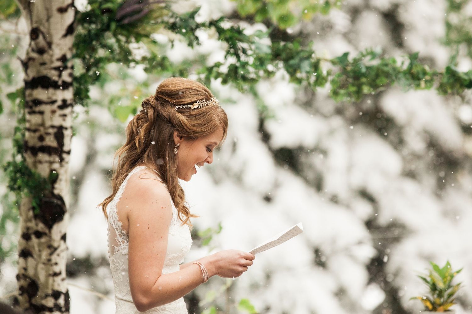 A bride in a white dress reads vows under tree branches during an outdoor wedding ceremony.